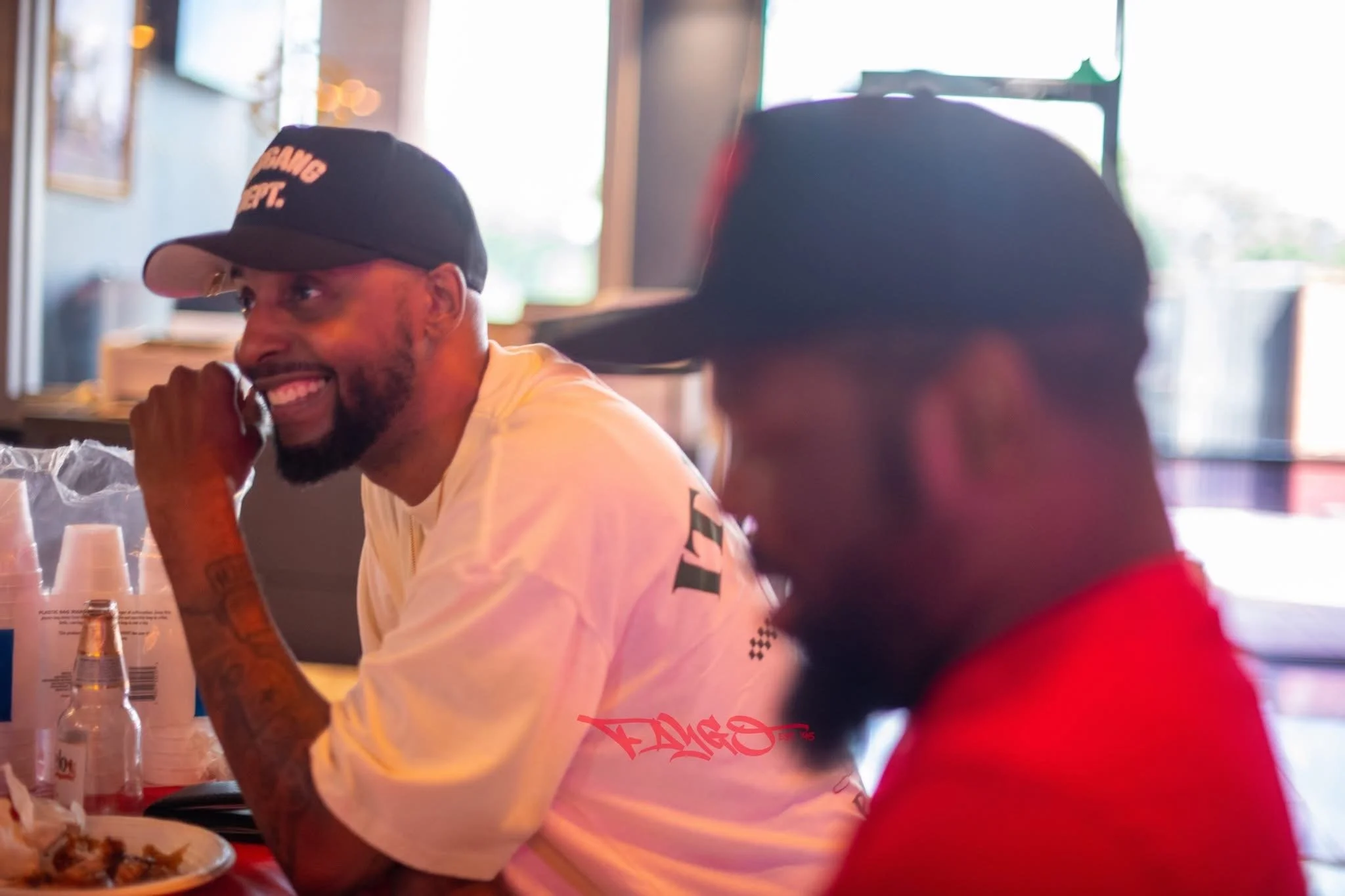 Two men seated at a table inside a restaurant, one wearing a black cap and white shirt, smiling and looking at the other, who is wearing a black cap and red shirt, with a cheerful atmosphere and sunlight streaming through windows.