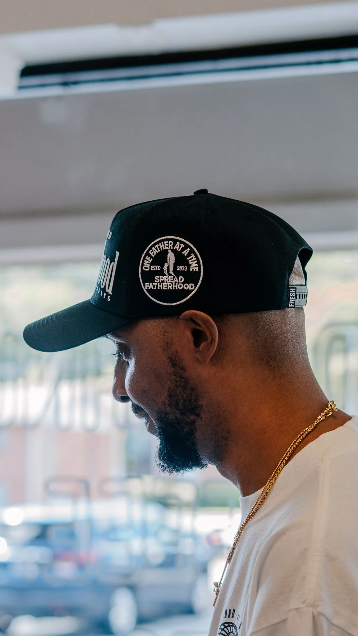 A man with a beard wearing a black baseball cap with a circular logo that reads 'One Father at a Time, Spread Fatherhood,' a white T-shirt, and a gold chain, standing indoors near a window.