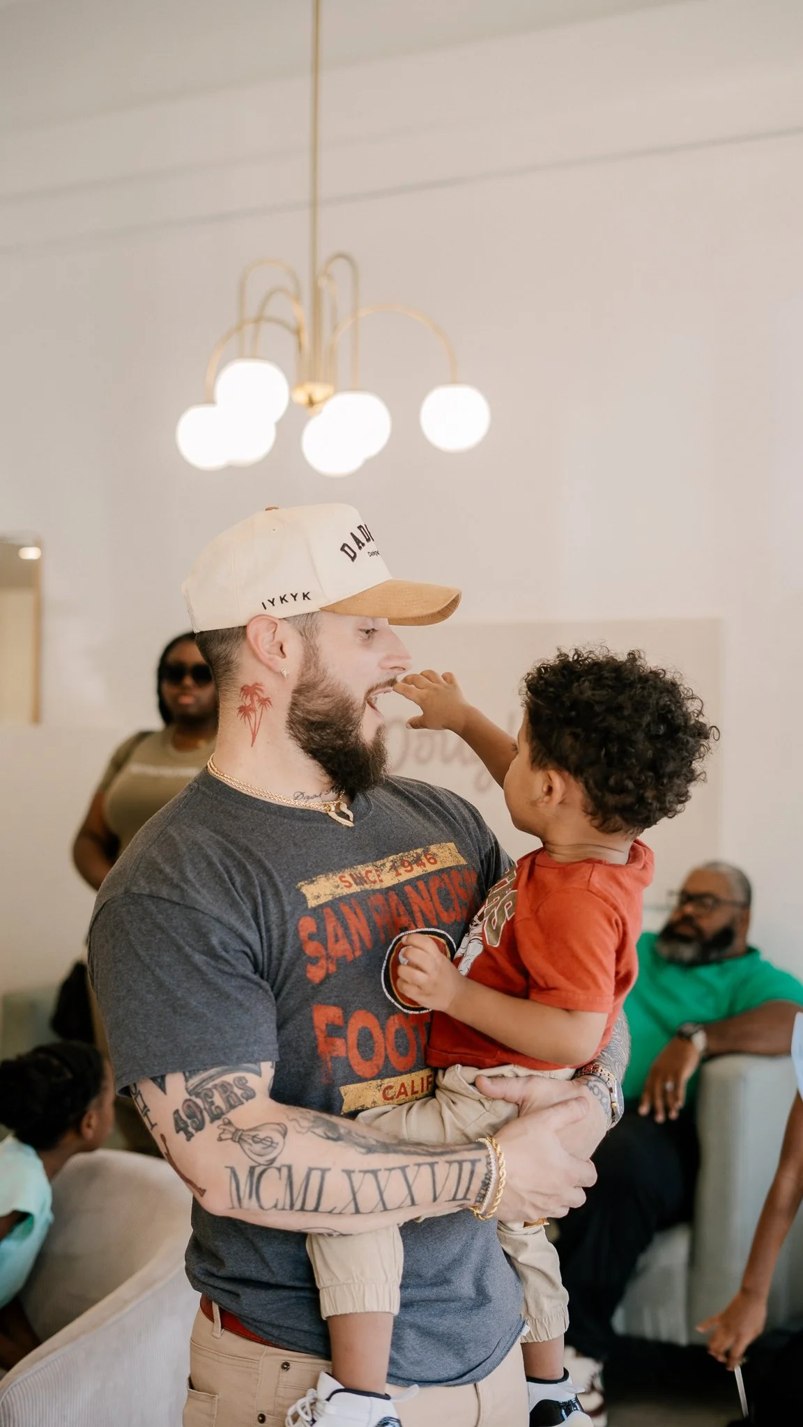 A man with tattoos and a beard holding a young child, touching his nose with the child's finger, in a living room with other people in the background.