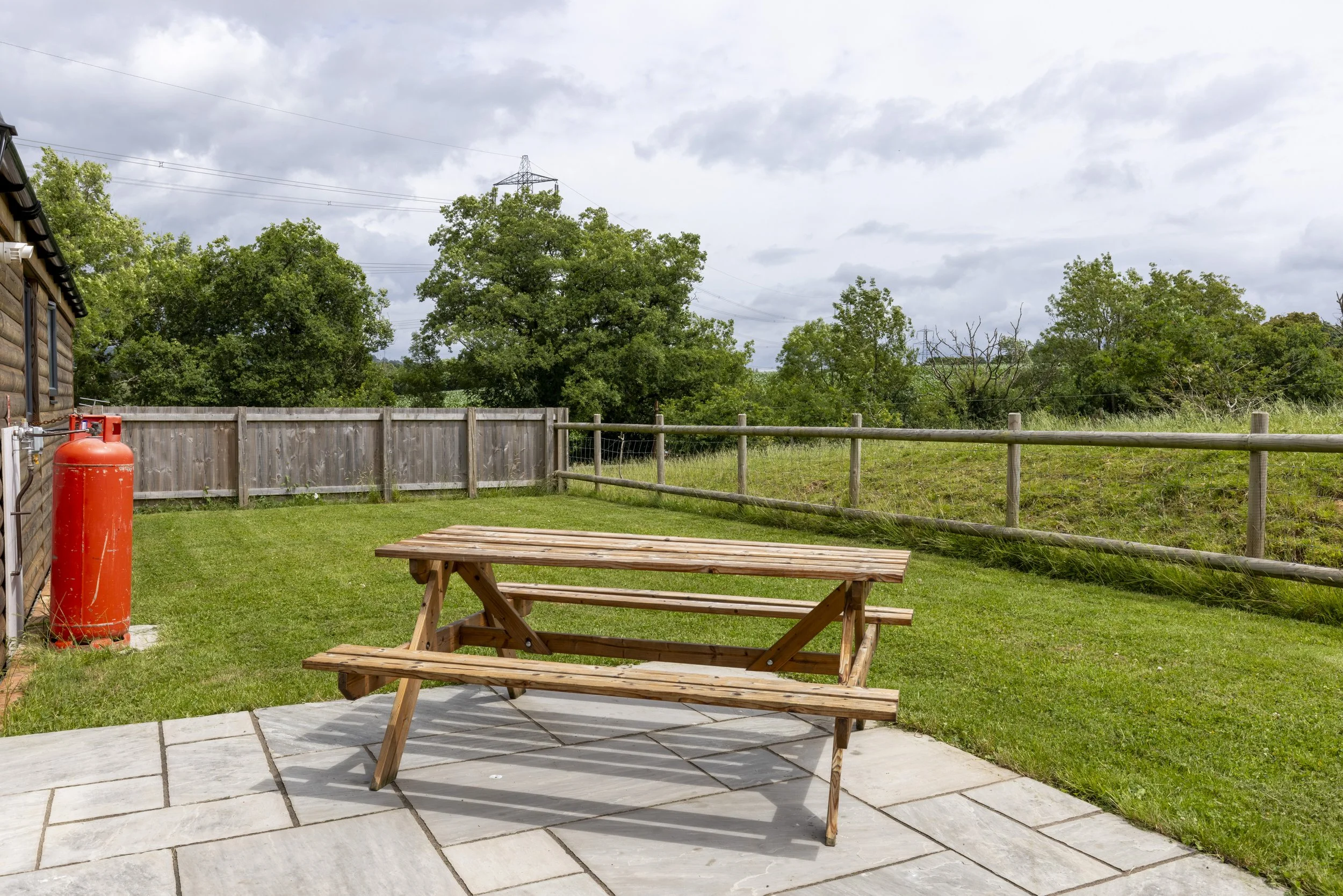 A backyard patio with a wooden picnic table on a stone tiled area, green grass, a wooden fence, trees, and cloudy sky in the background.