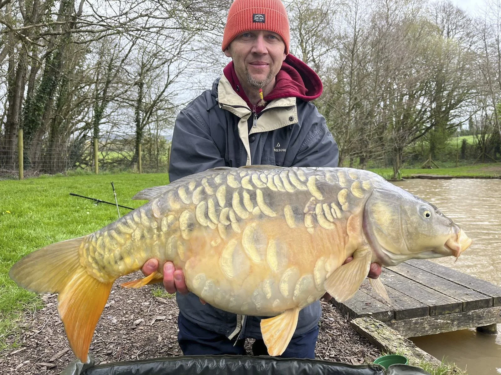A man in outdoor clothing holding a large carp fish near a pond.