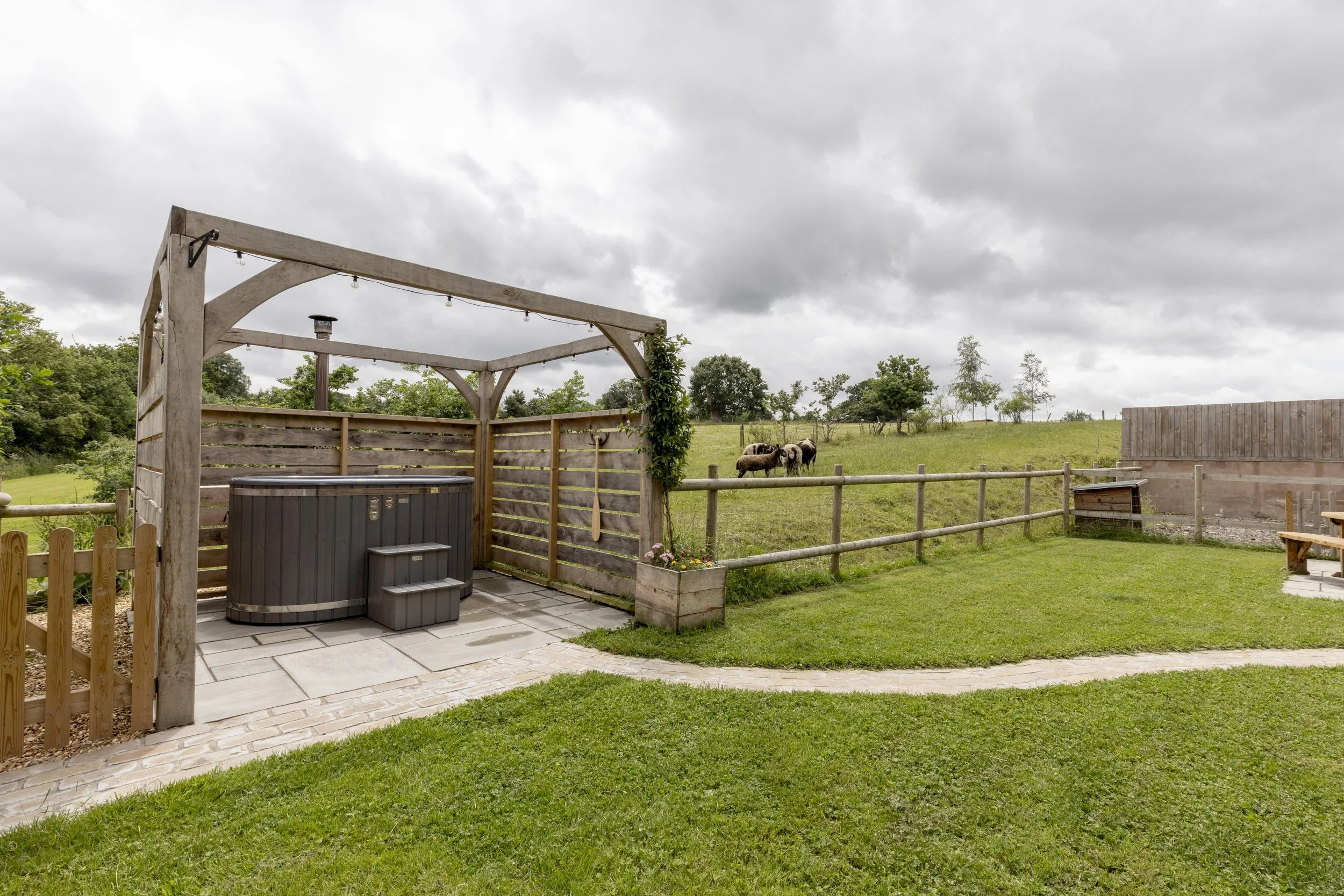 A fenced backyard with a small patio area, hot tub, and a wooden pergola, overlooking a grassy field with a herd of sheep grazing under a cloudy sky.