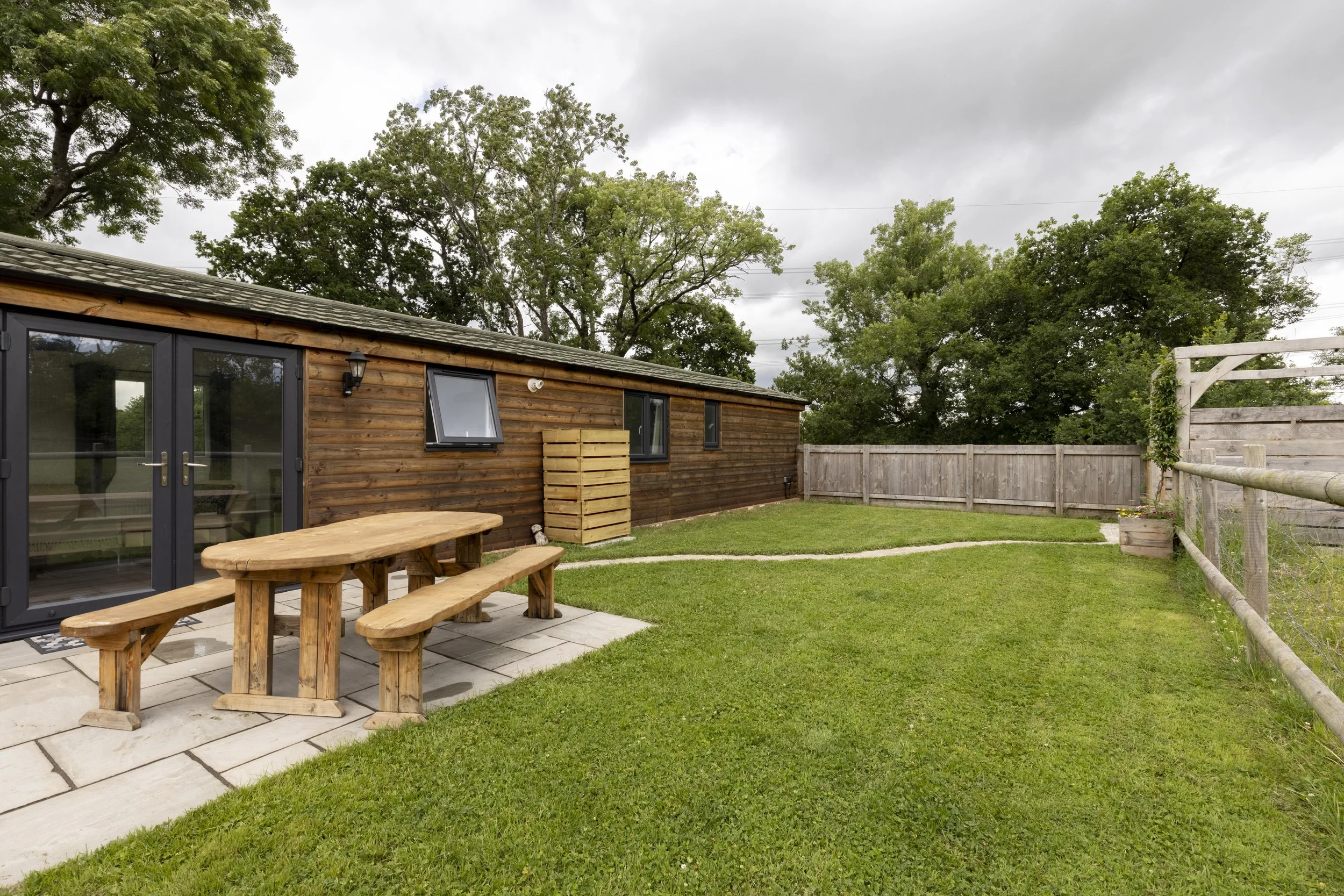 A backyard with a wooden shed, picnic table, grassy lawn, and a wooden fence, under a cloudy sky with tall trees in the background.