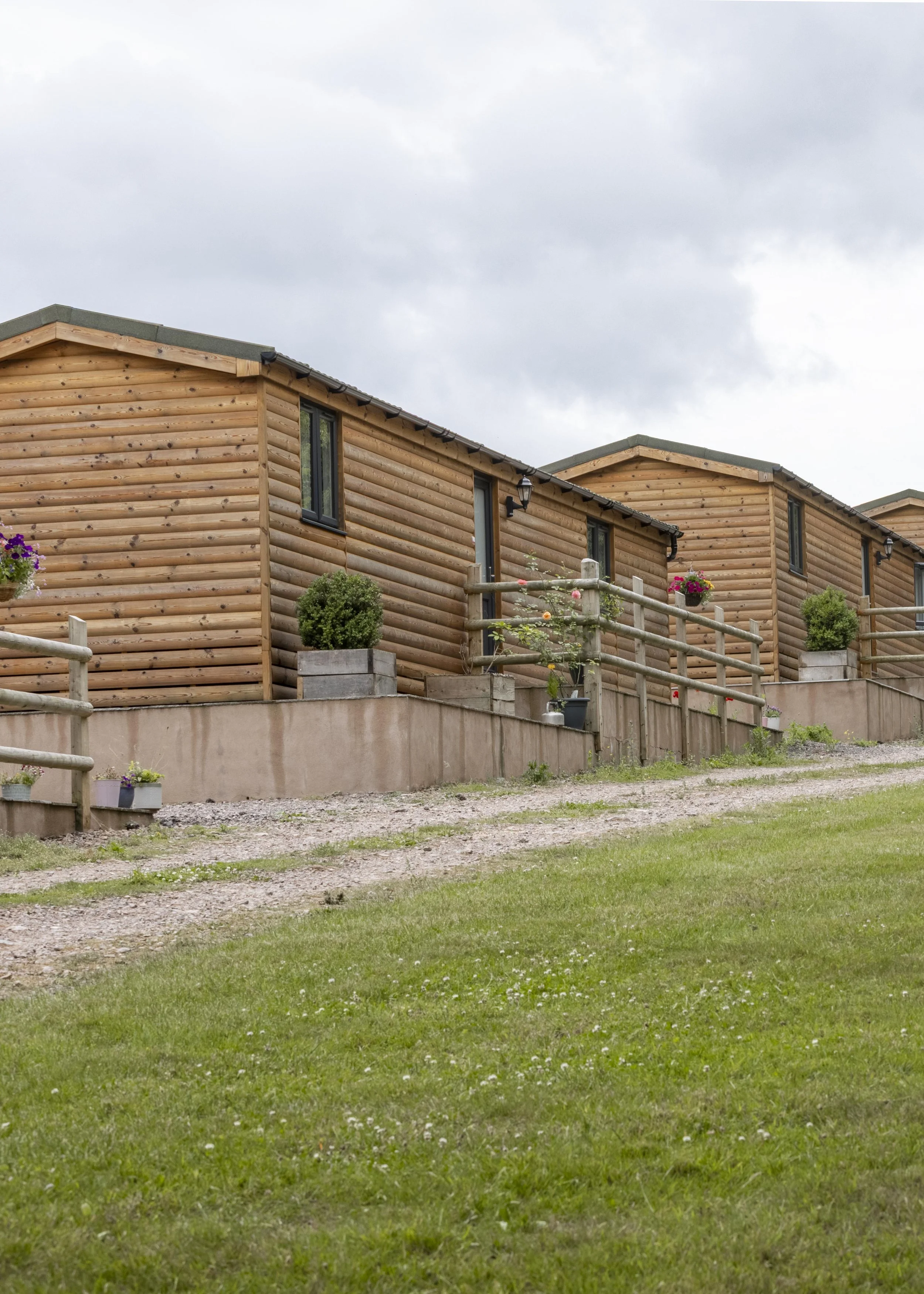 Wooden houses with black window frames and flower pots, surrounded by a wooden fence and green grass, under a cloudy sky.