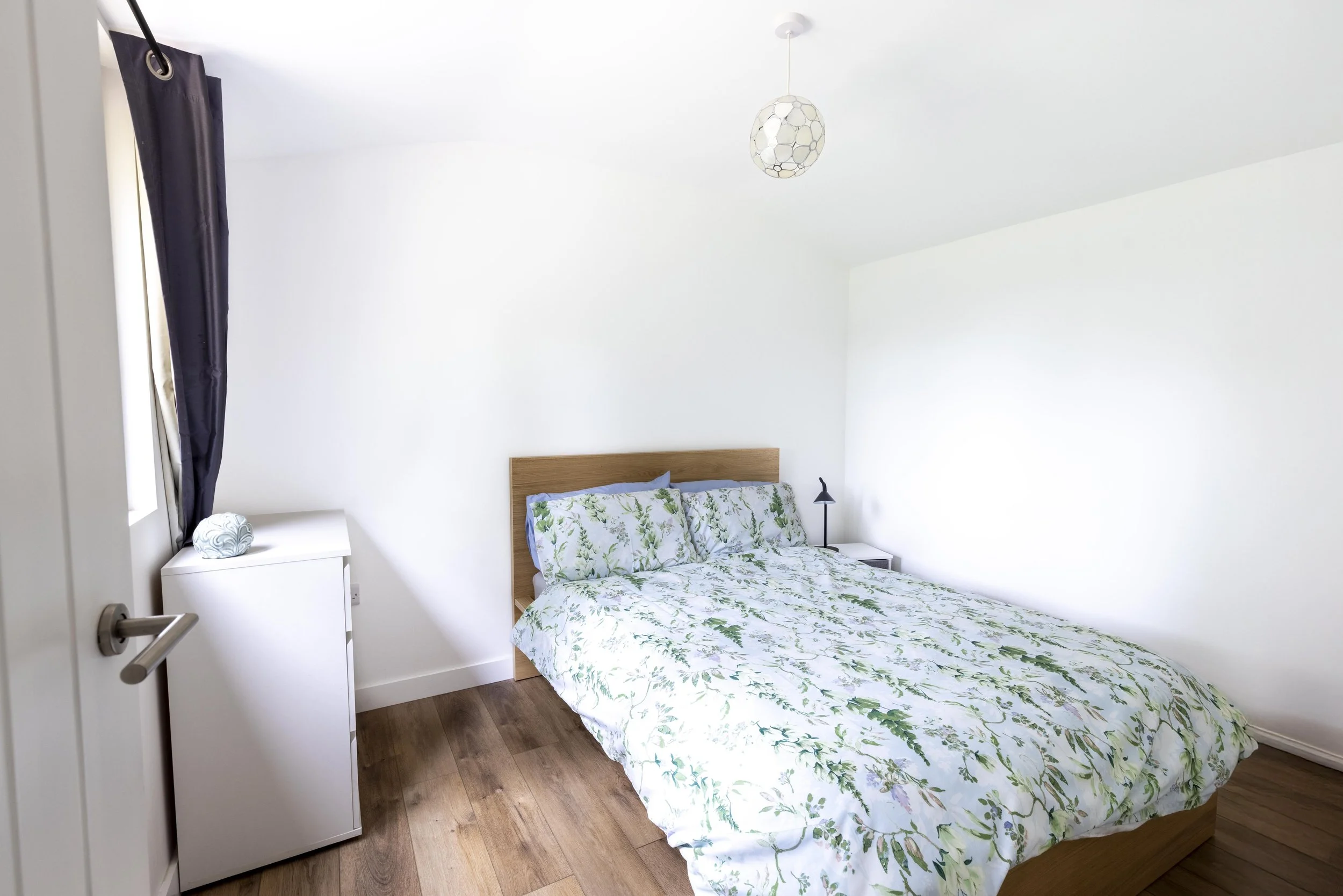 A bedroom with a bed dressed in a white and green leafy patterned duvet and pillows, a small white nightstand with a decorative orb, a black desk lamp, a dark curtain, and a white ceiling with a geometric pendant light.