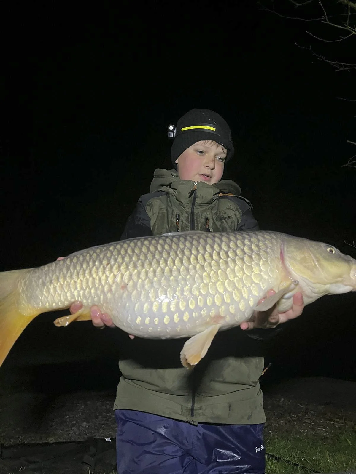 A young boy at night holding a large fish with both hands, wearing a black hat with a headlamp, green jacket, and blue waterproof pants.