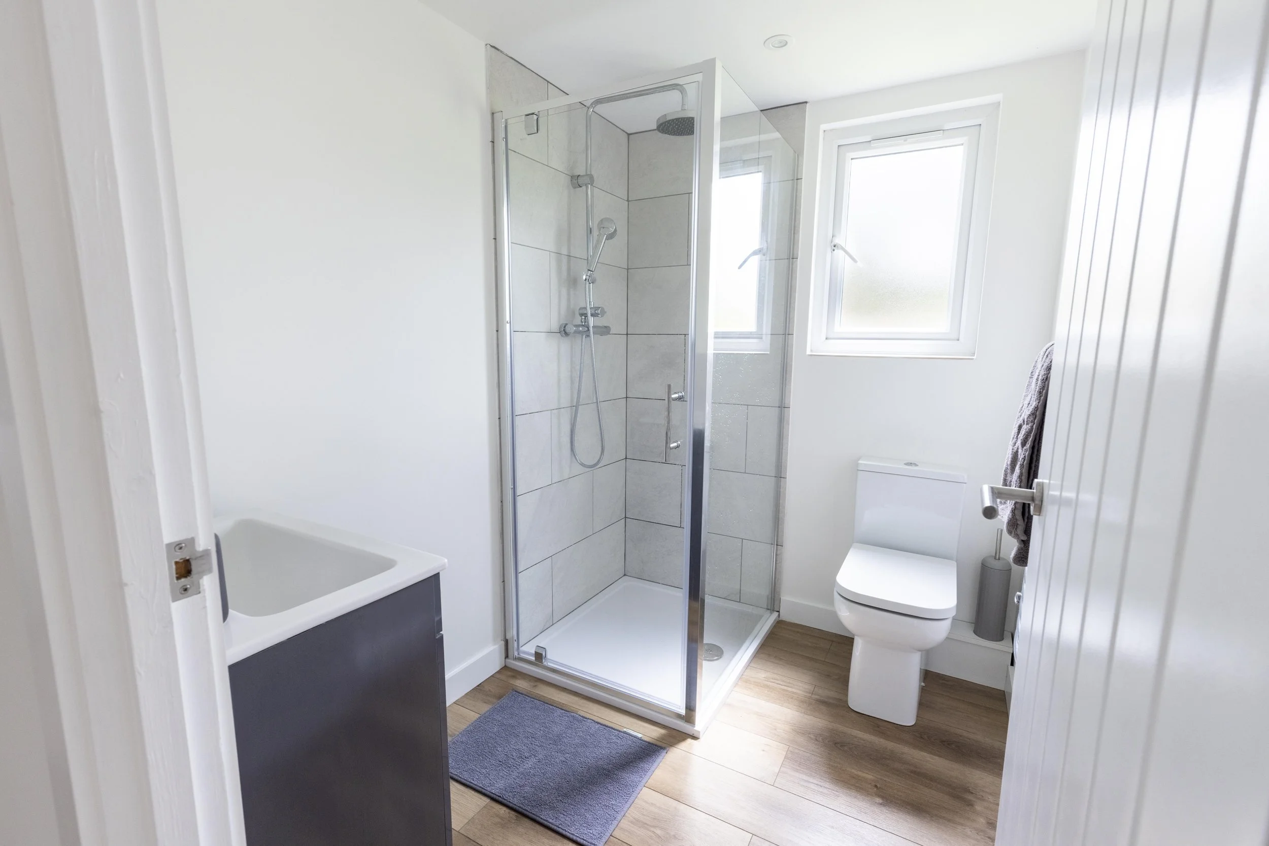 A modern bathroom with white walls, a glass-enclosed shower with tiled walls, a toilet, a bathtub, and a window letting in natural light.