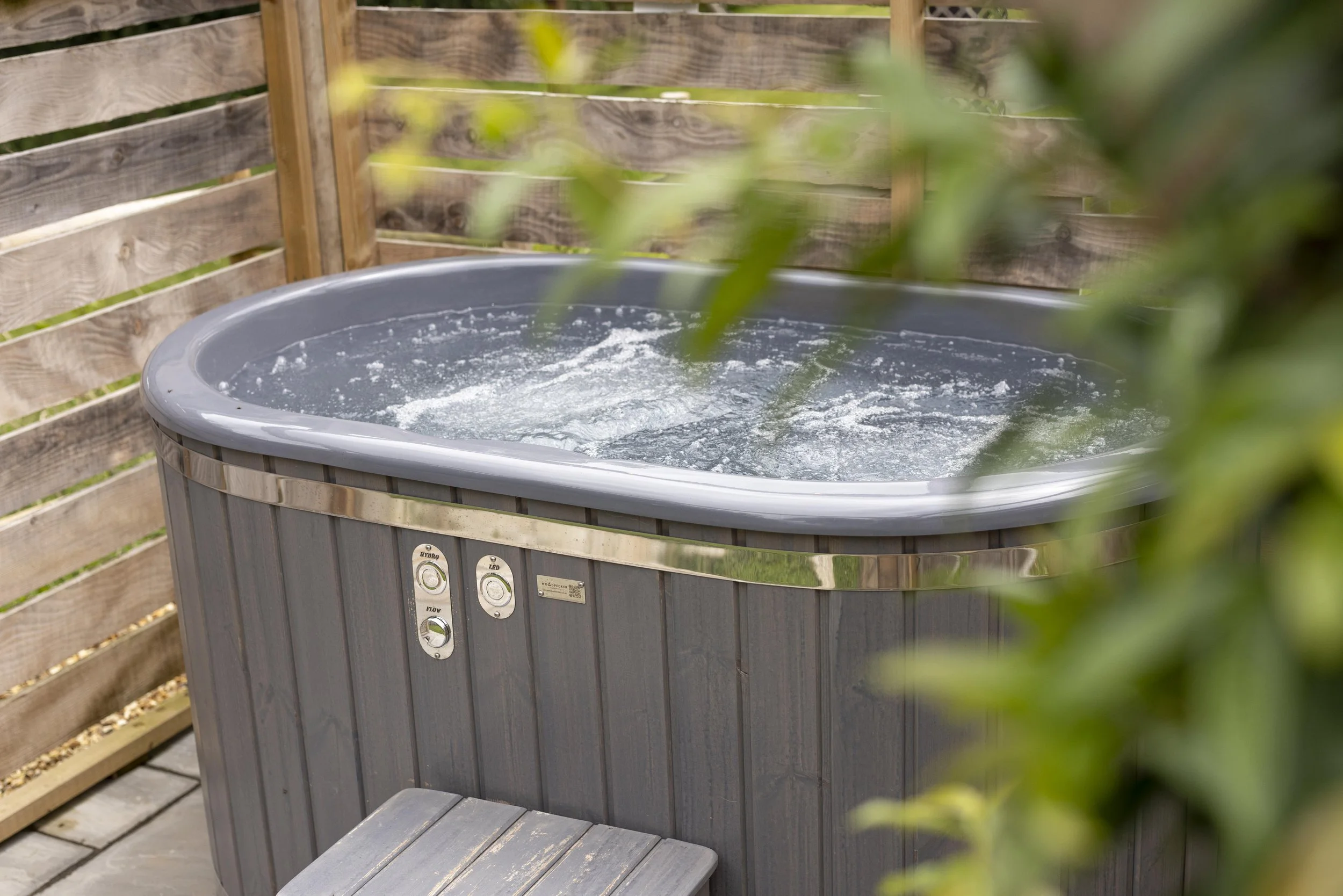 A hot tub on a wooden deck against a wooden fence, with a small wooden bench in front and some green leaves partially obscuring the view.