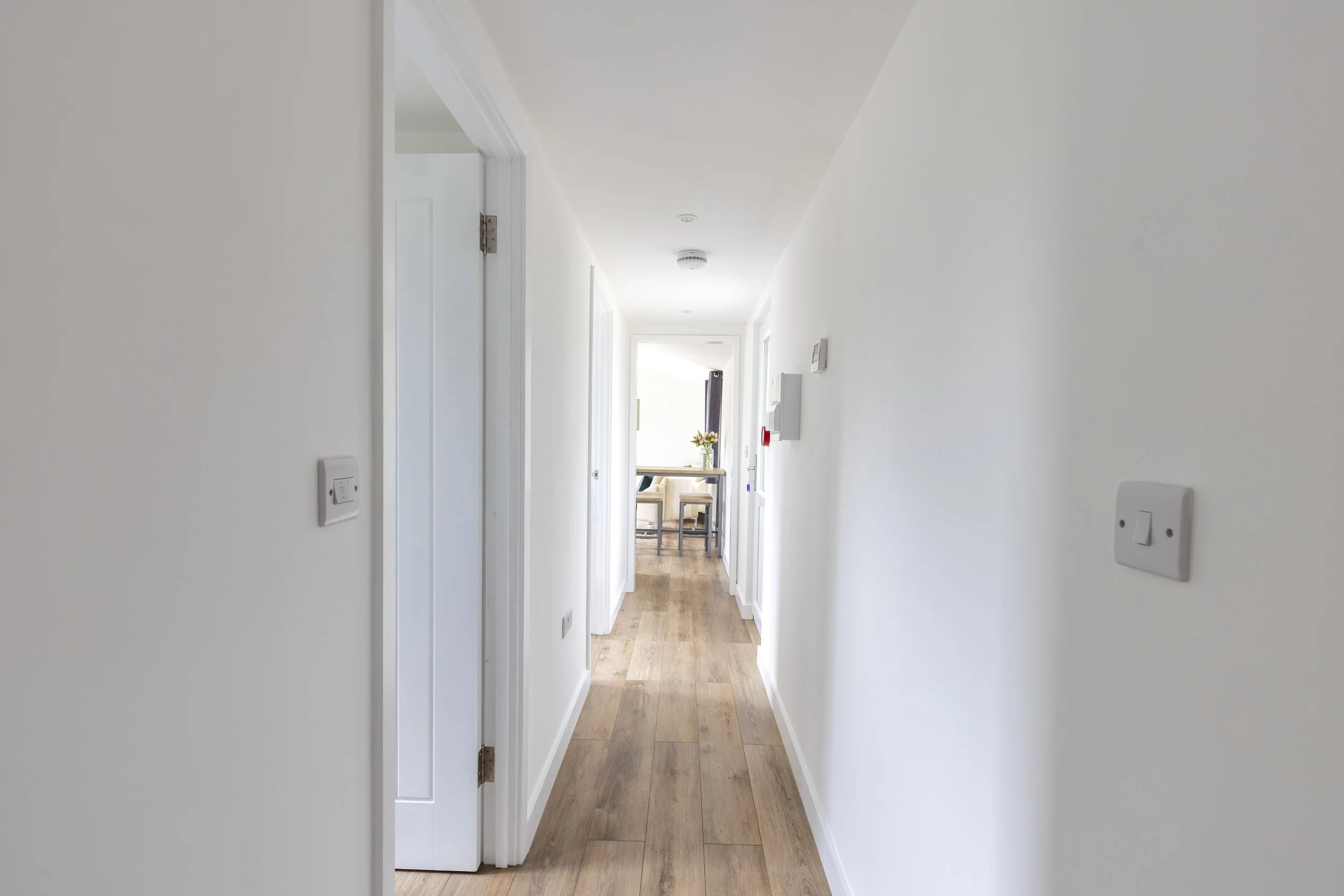 A bright, narrow hallway with white walls, wooden flooring, and a door at the end with a windows and curtains, a table with a vase of flowers is visible through the door.