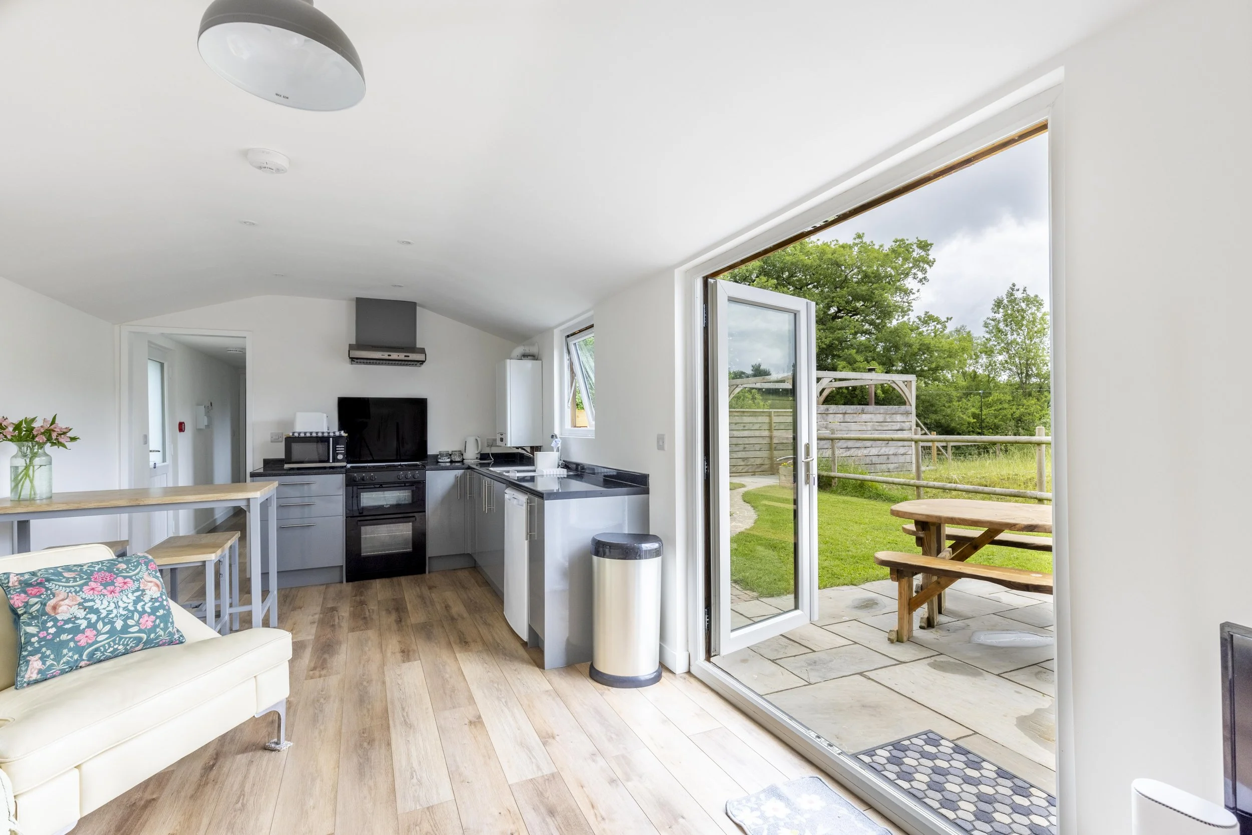 Interior view of a modern kitchen with a door leading outside to a patio and grassy yard with trees, a picnic table, and a wooden fence.