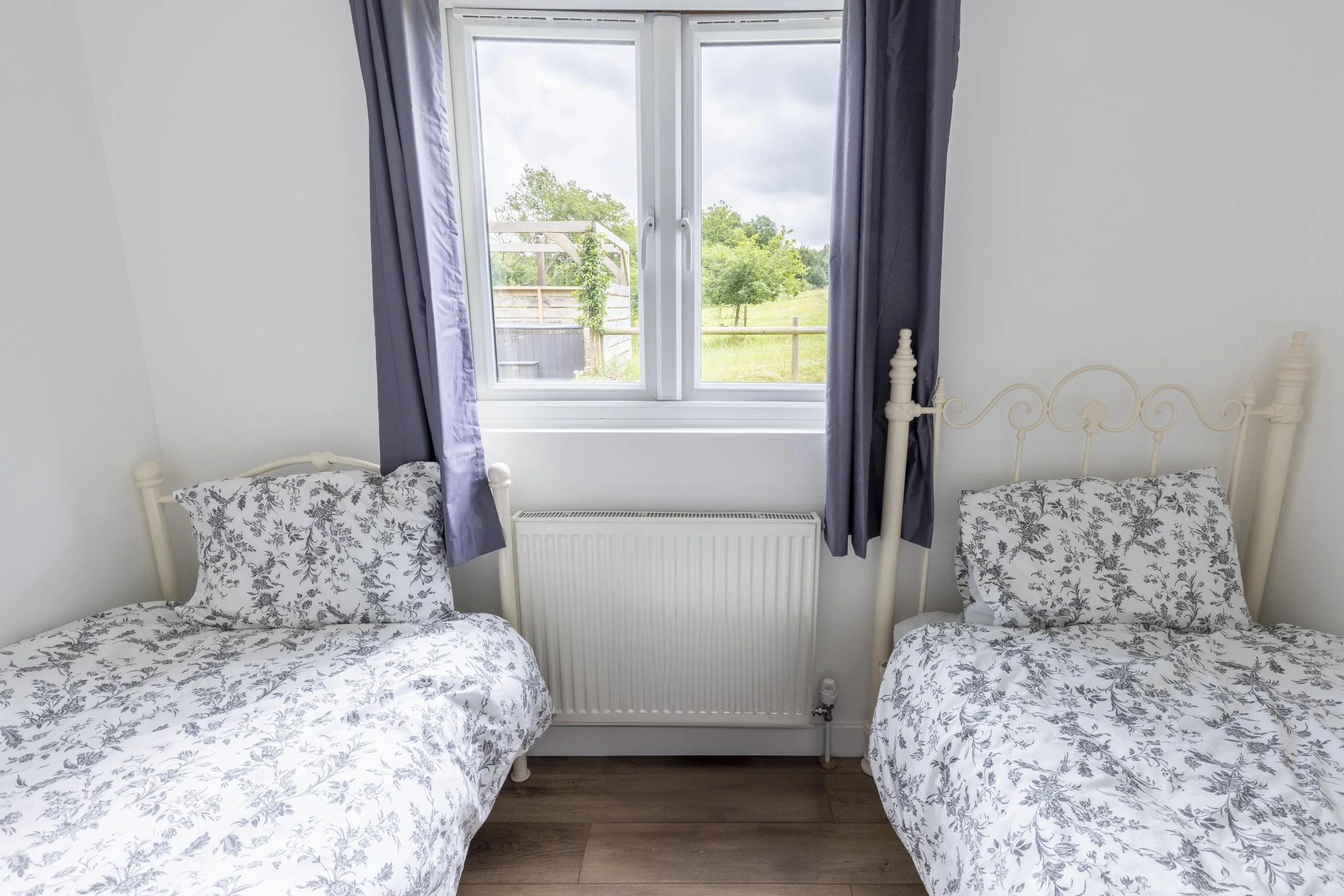 Two beds with matching floral bedspreads and pillows in a room with a large window showing an outdoor view of trees and a grassy field.