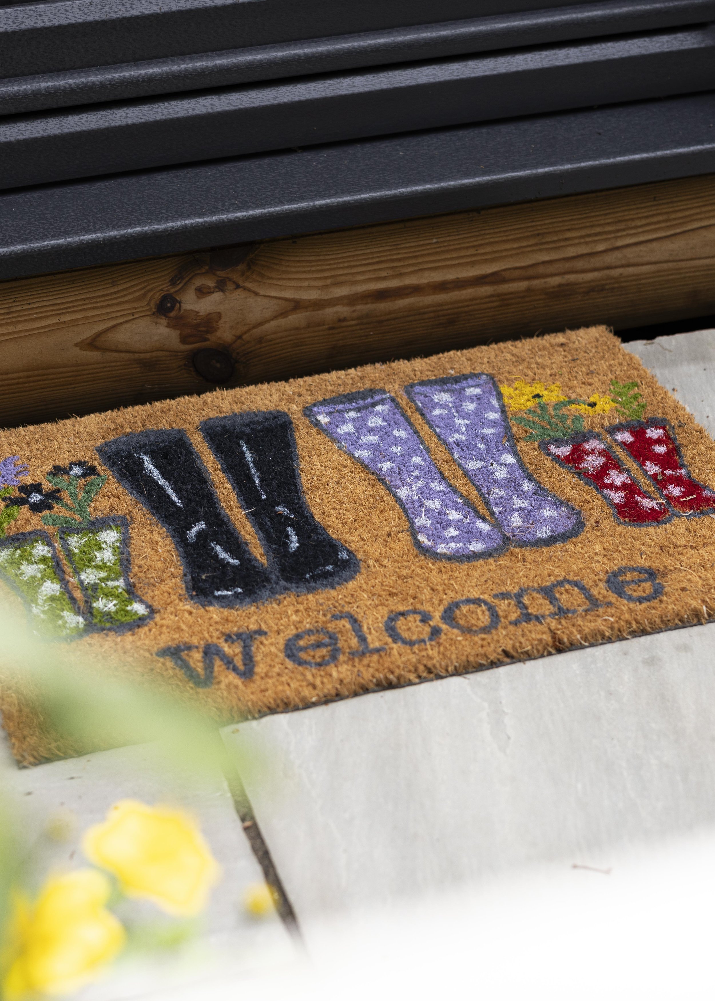 A doormat with colorful flower designs and the word 'WELCOME' written in black letters. The doormat is placed outside near a wooden step with black matting on top.