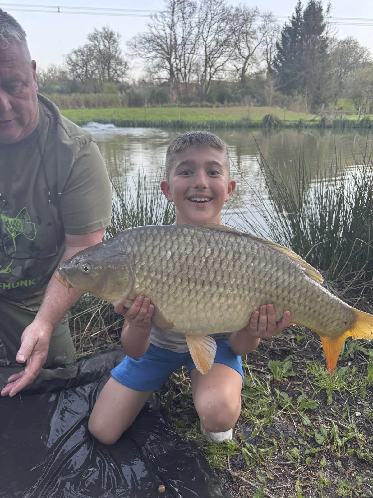 A young boy holding a large fish, smiling at the camera, next to an adult man by a river with trees in the background.