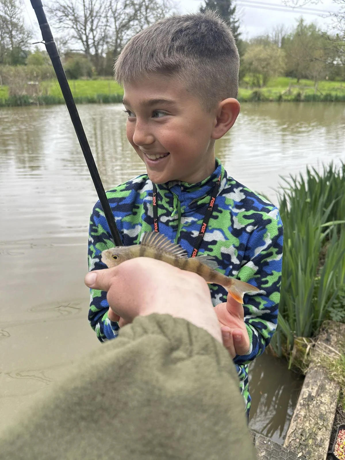 A young boy with short hair smiling as he holds a small fish caught while fishing near a lake or pond, with trees and grassy area in the background.