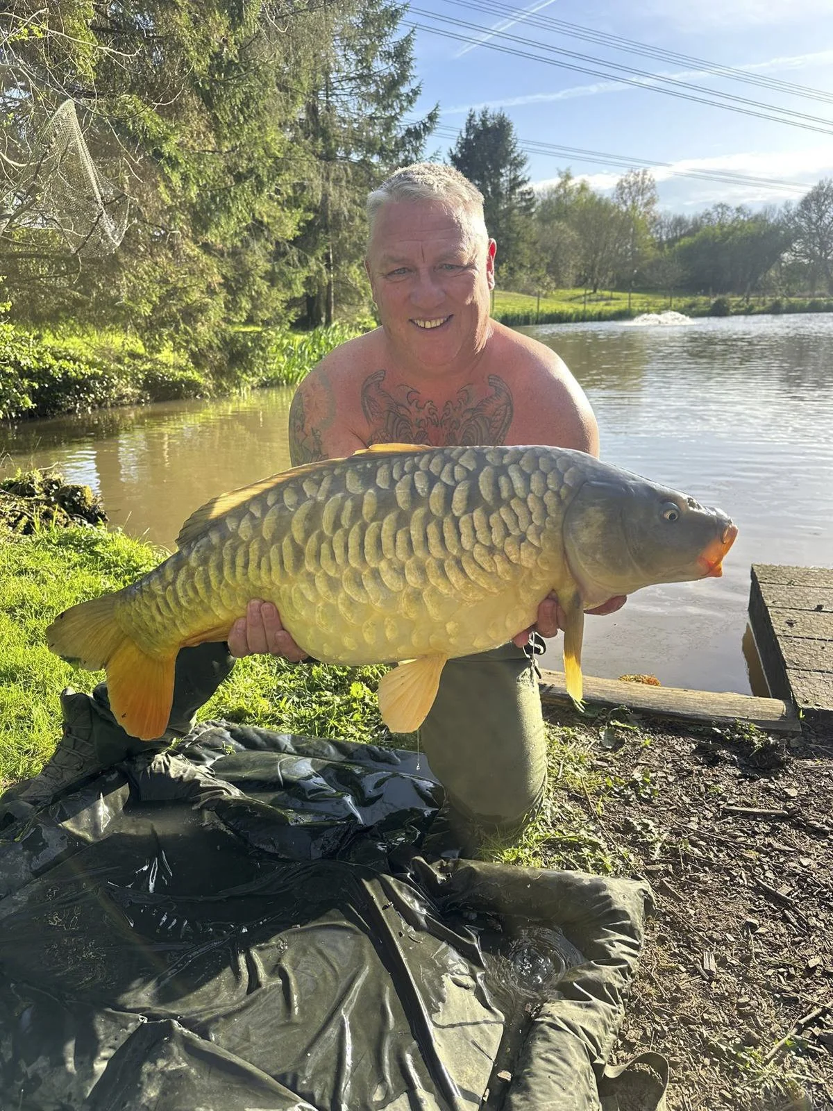A man holding a large fish near a riverbank with trees and blue sky in the background.