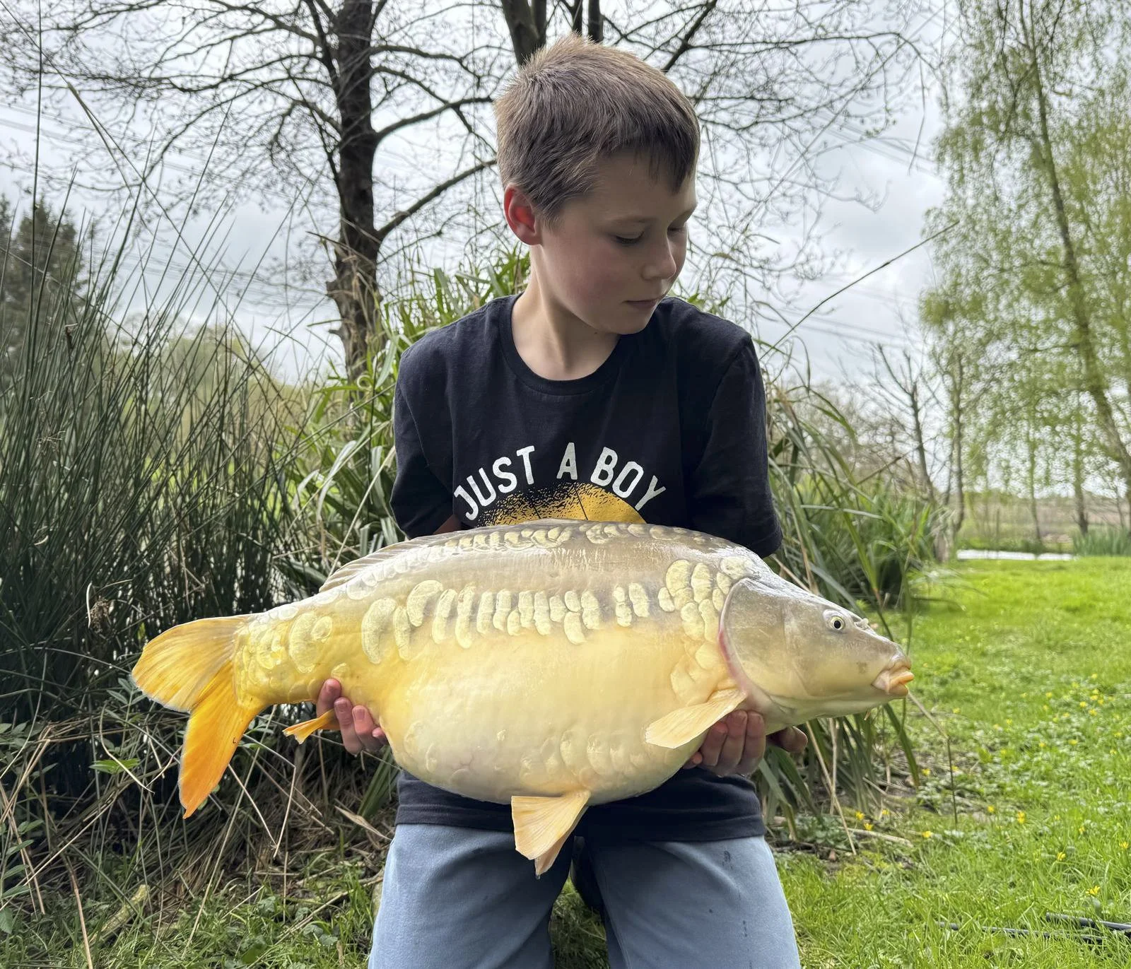 A young boy holding a large yellow fish outdoors surrounded by greenery and leafless trees.