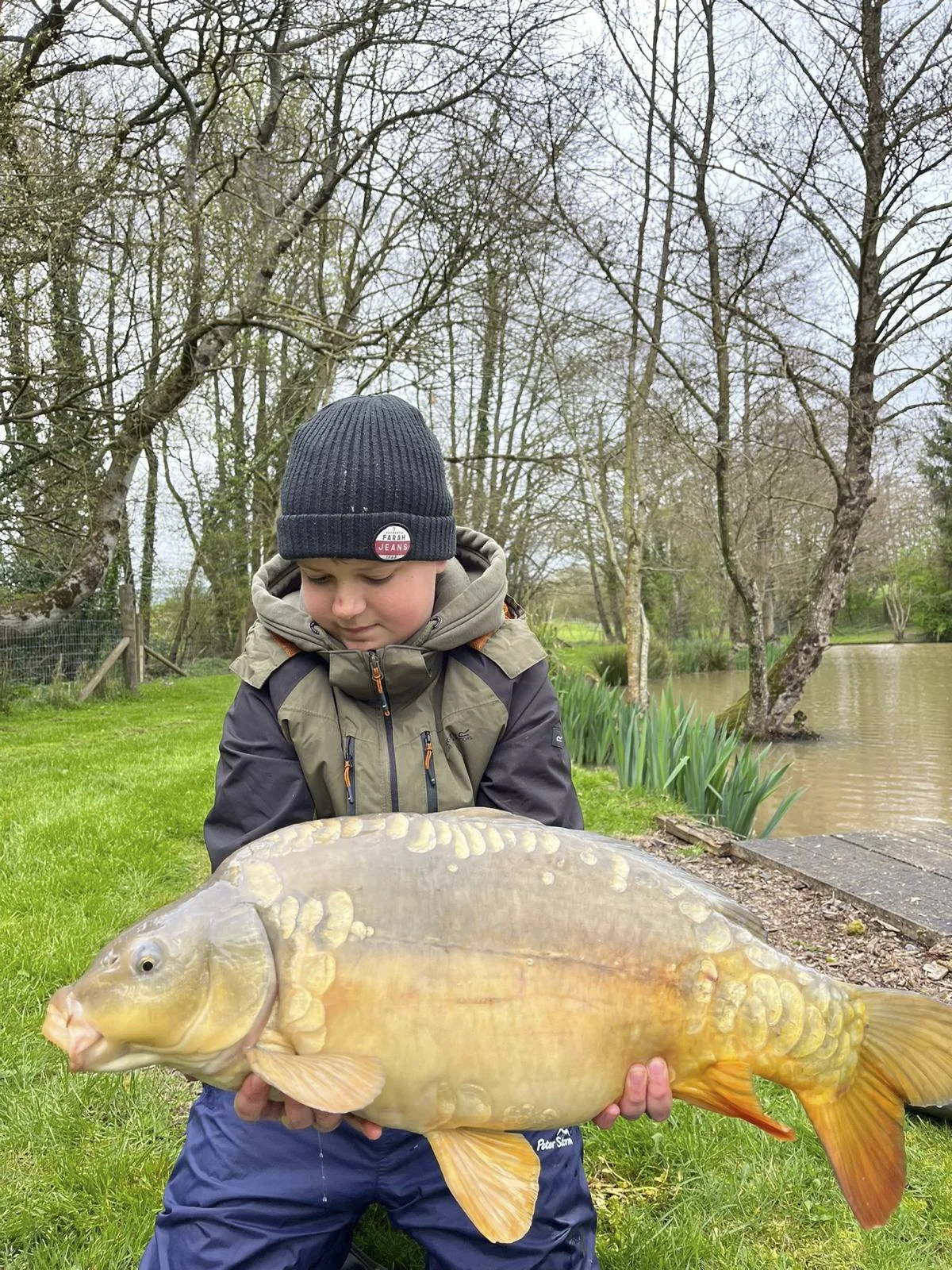 A boy in a beige and black waterproof jacket and a black beanie hat holding a large fish outdoors next to a pond with bare trees.