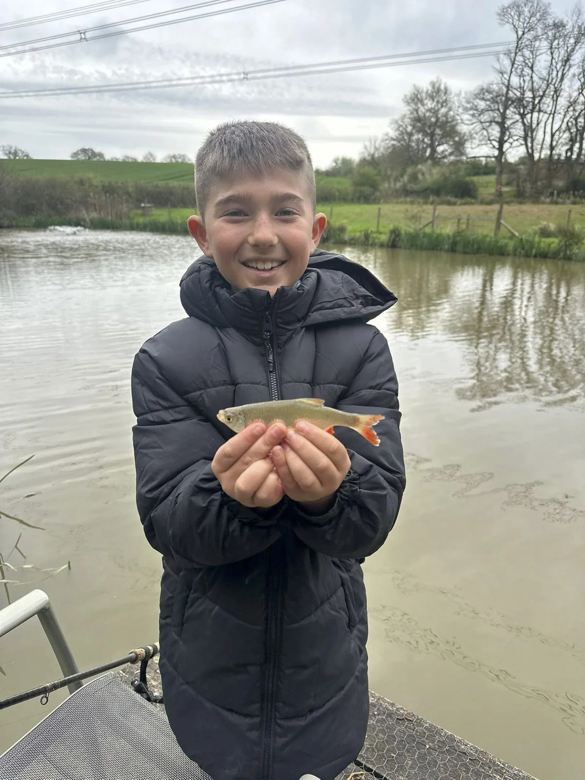 A young boy standing on a boat by a lake, holding a small fish, smiling at the camera, with trees and overcast sky in the background.