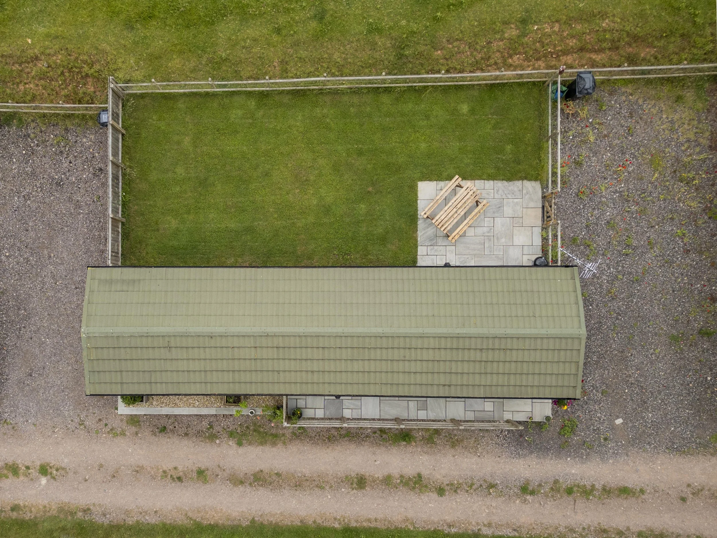 An aerial view of a backyard divided into a grassy section and a gravel area, with a small patio and wooden pallet on the grass, a garden area with plants, and a fence surrounding the yard.