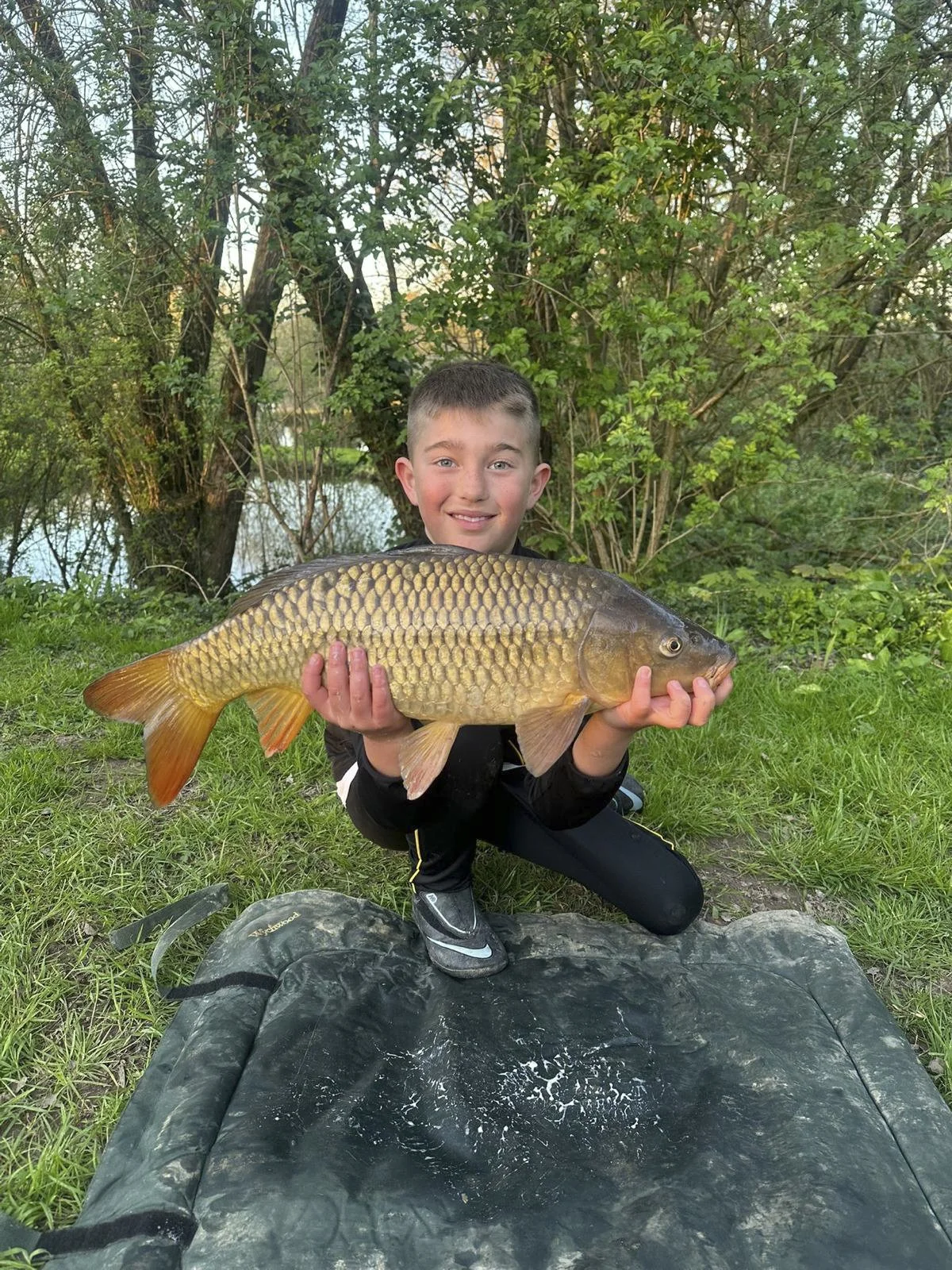 A young boy kneels on grass, smiling, and holds a large fish horizontally in front of him with both hands. The fish has golden scales and orange fins. Behind him are trees and a body of water, suggesting an outdoor fishing setting.
