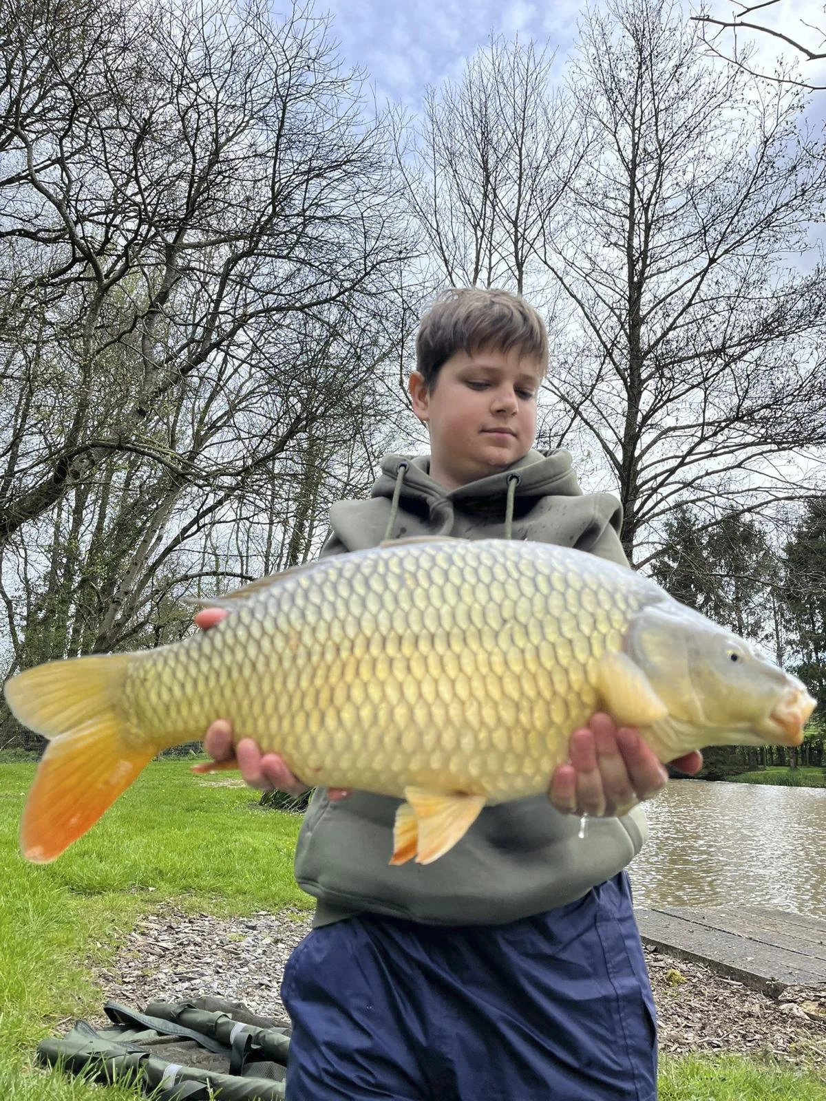 A young boy holding a large, yellowish fish with orange fins, outdoors near a river with trees and grass in the background.