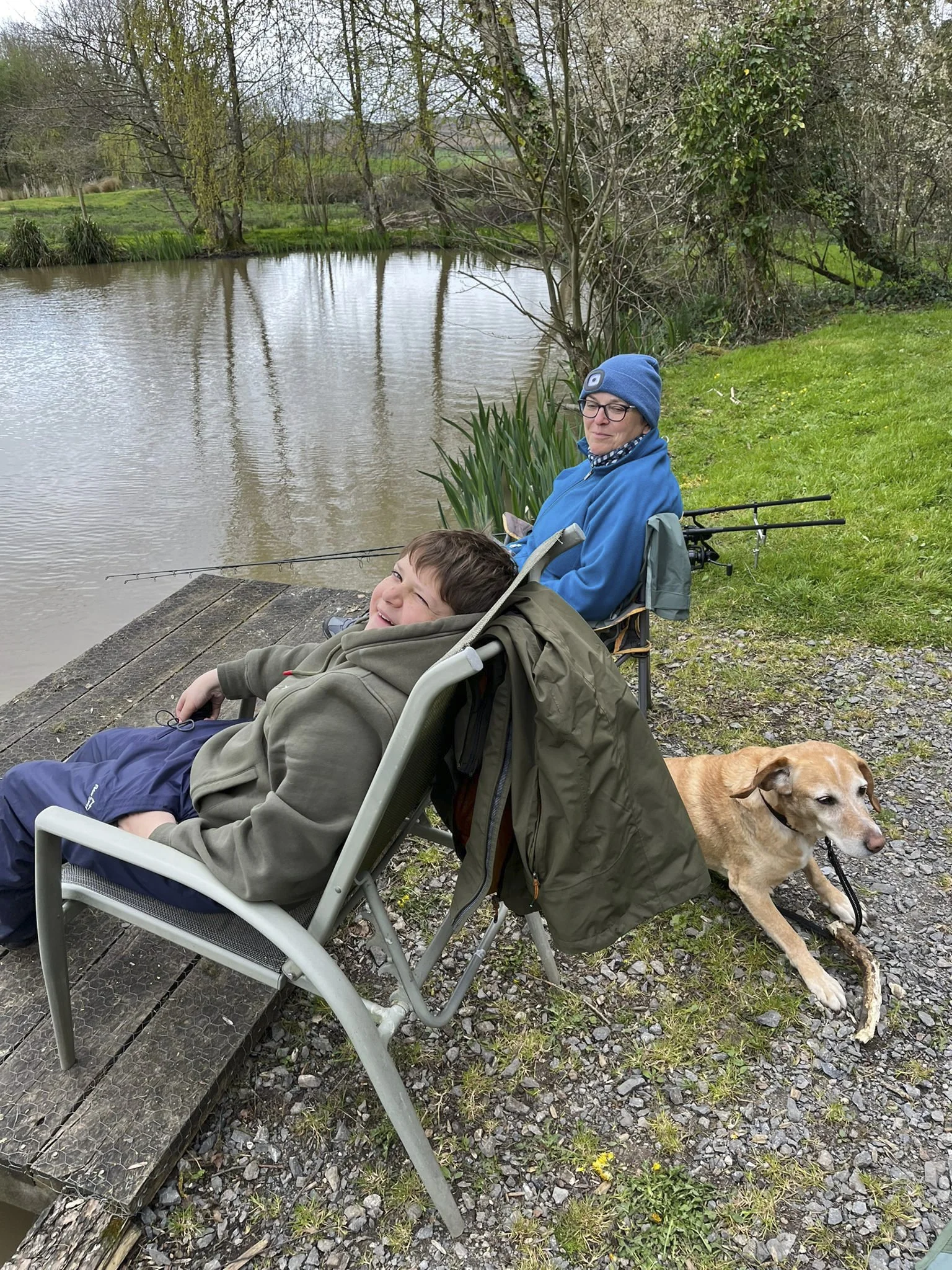 A woman and a young boy sitting in chairs by a pond, with a dog lying on the grass nearby, surrounded by trees and grassy areas.