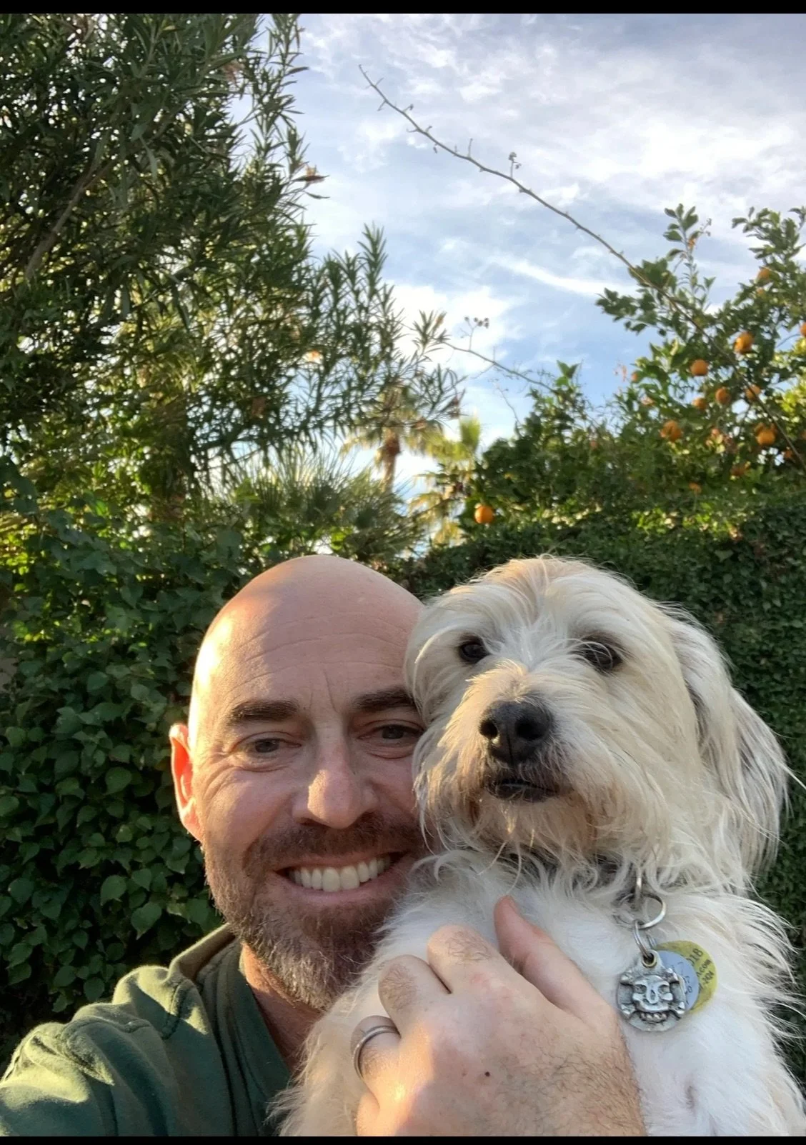 A man smiling while holding a small, fluffy dog outdoors with lush green trees and a blue sky in the background.