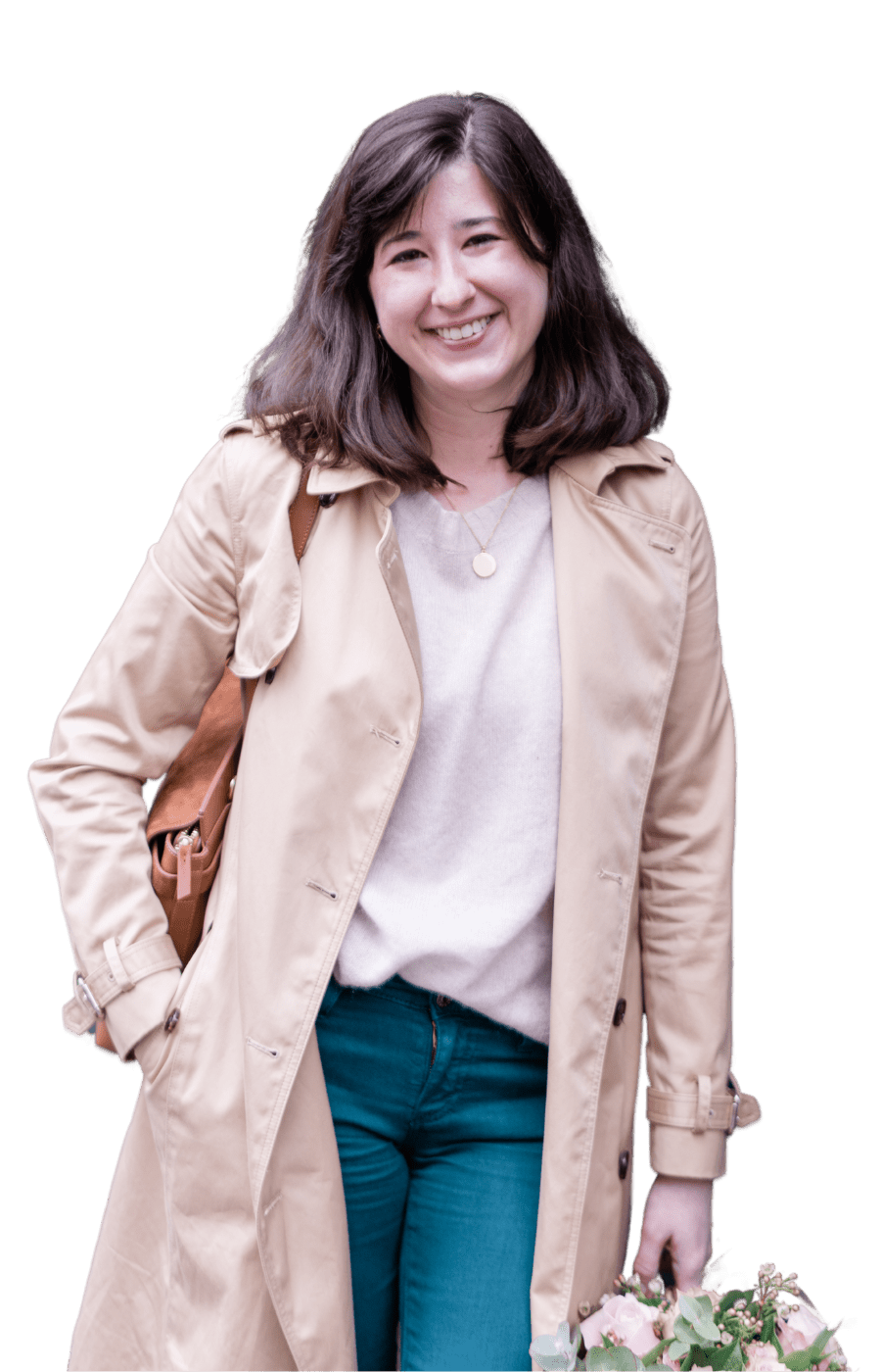 A smiling woman with shoulder-length brown hair, wearing a beige trench coat, a light gray sweater, and blue jeans, carrying a brown shoulder bag and holding a bouquet of flowers.