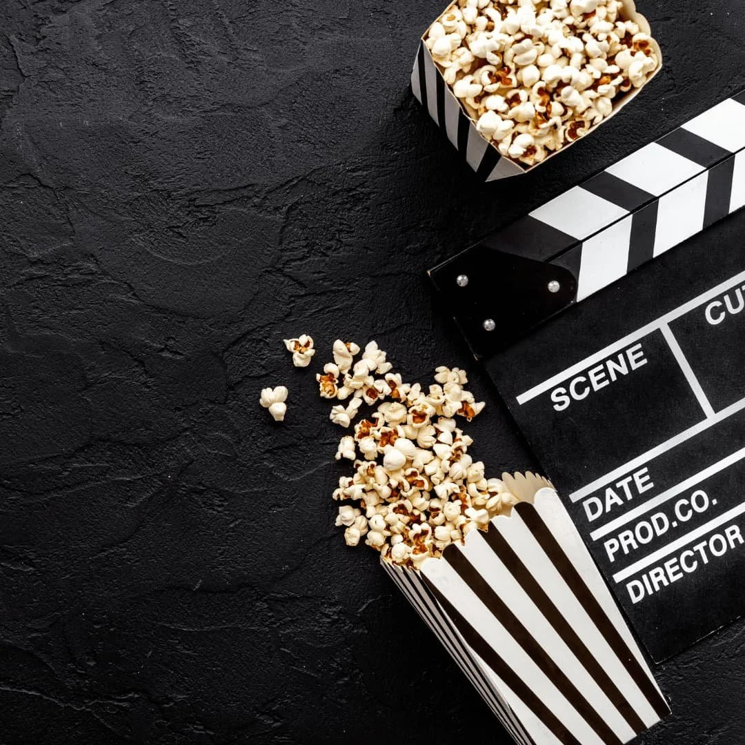 Popcorn in a black and white striped container spilled on a black textured surface with a black and white clapperboard nearby.