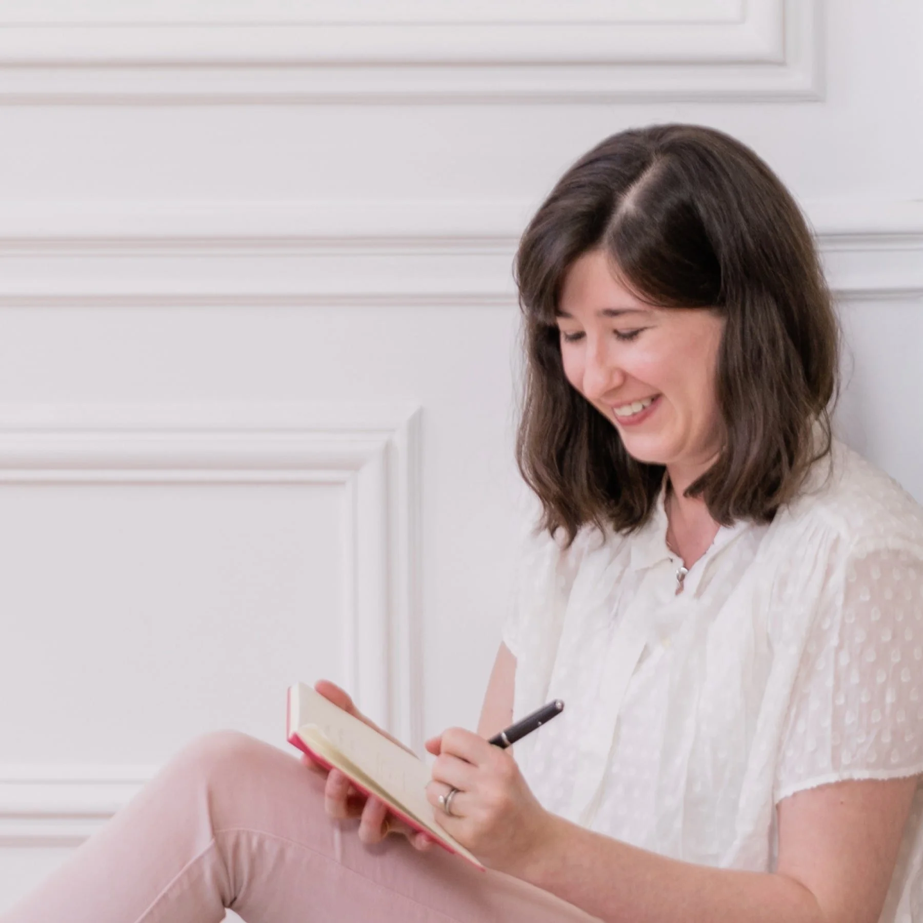A woman with shoulder-length brown hair is sitting against a white paneled wall, smiling while writing in a small notebook with a black pen.