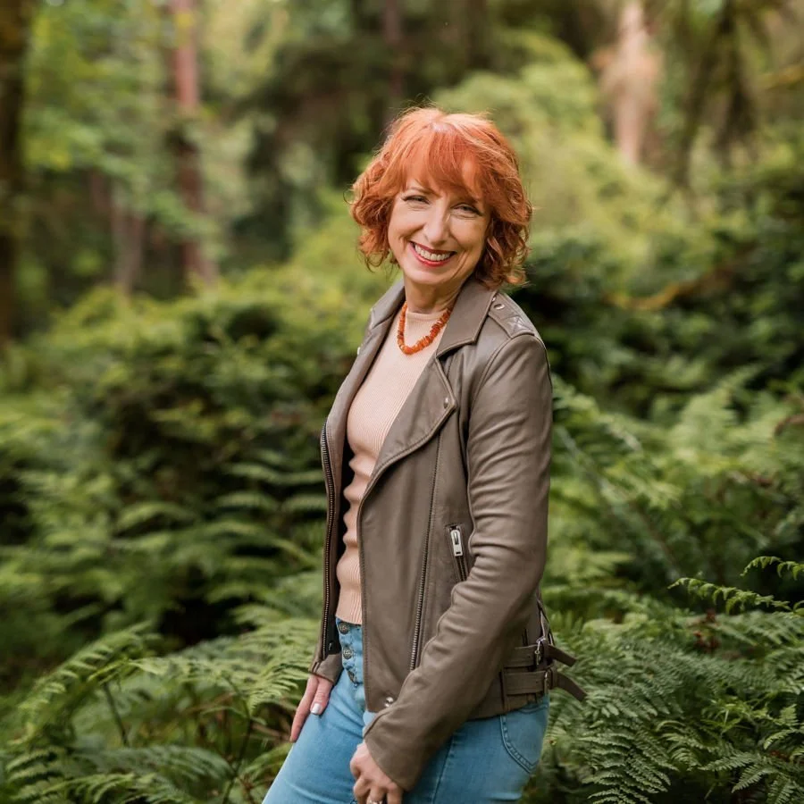 A woman with red curly hair smiling outdoors in a lush green forest, wearing a beige top, gray leather jacket, and blue jeans.