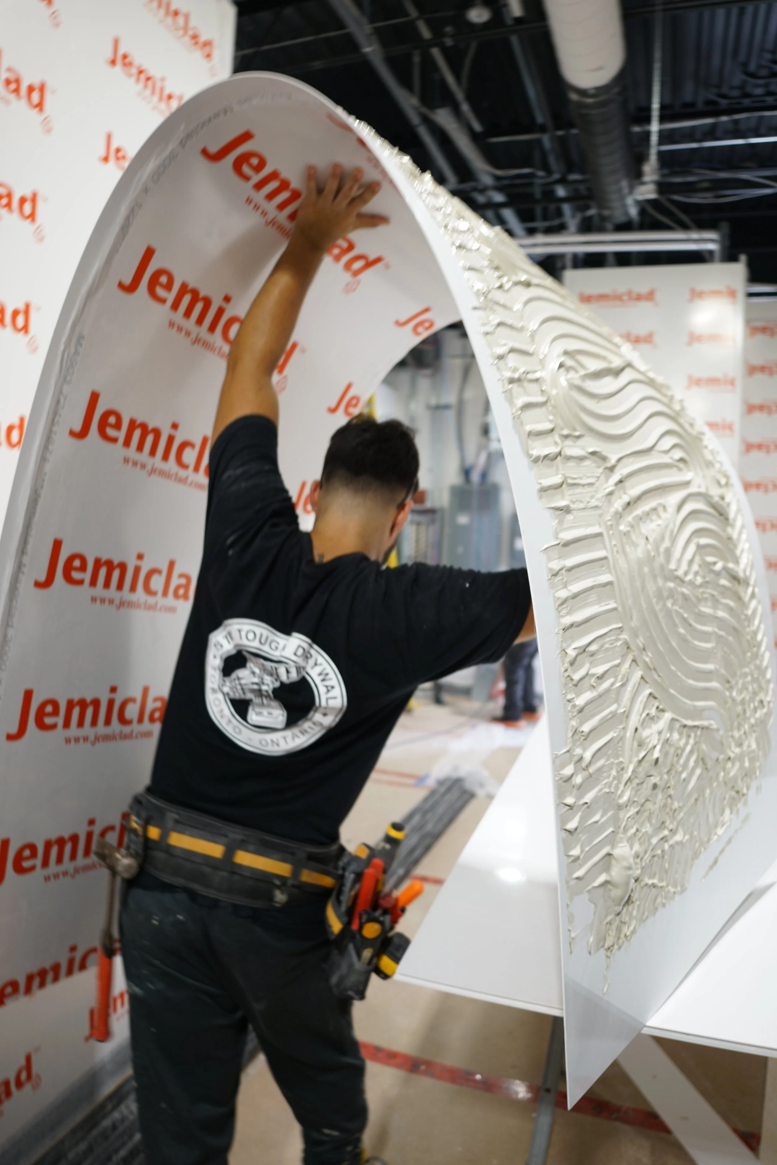 A worker with a tool belt applies textured white material to a large curved white panel inside a construction site or showroom.