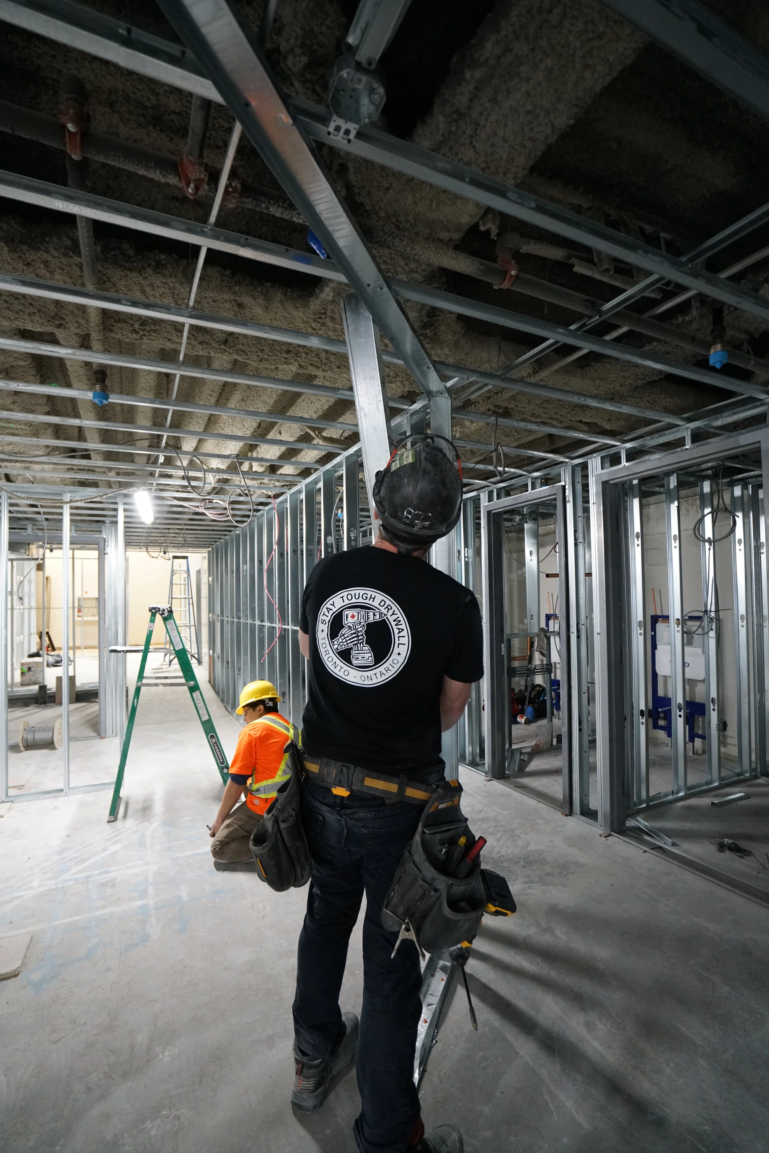 Two construction workers in a building under construction, with exposed metal framing and electrical wiring. One worker, wearing a black shirt and helmet, is looking up at the ceiling, while the other, wearing orange safety gear and a yellow helmet, 