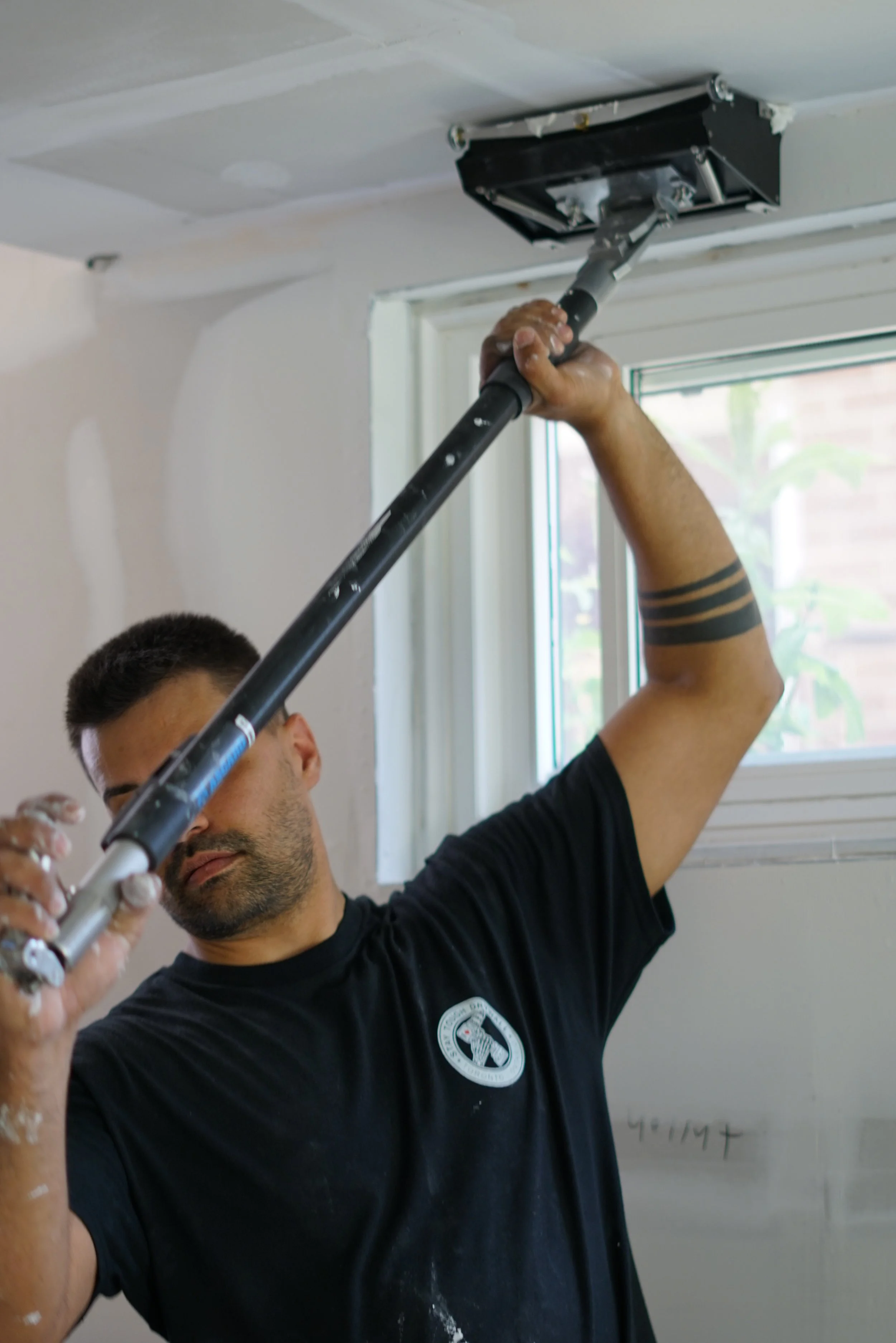 Man painting a ceiling with a roller extension pole near a window.
