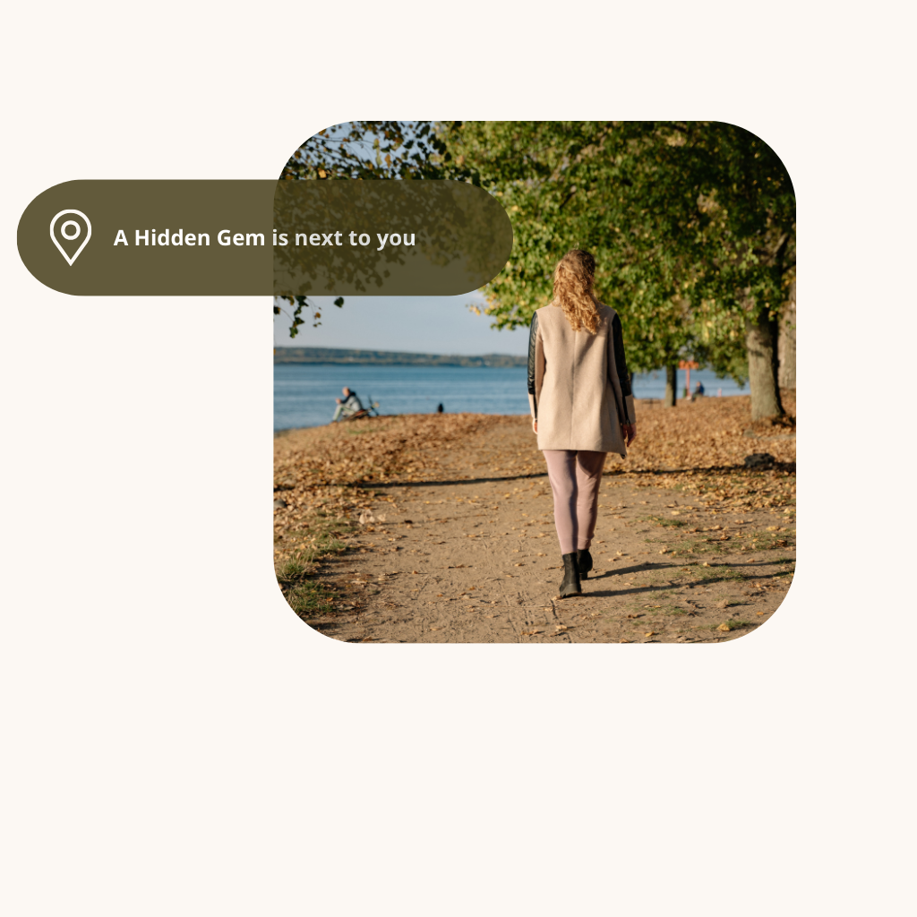 A woman walking along a dirt path near water with trees and a person sitting on a bench in the background