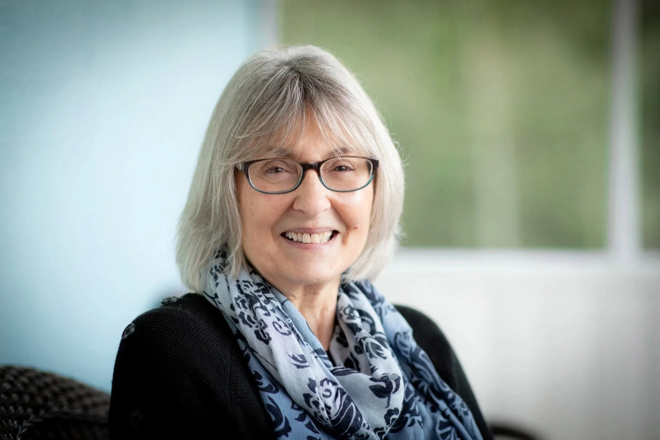 Older woman with gray hair, black glasses, smiling, wearing a scarf and black top, indoors near window.
