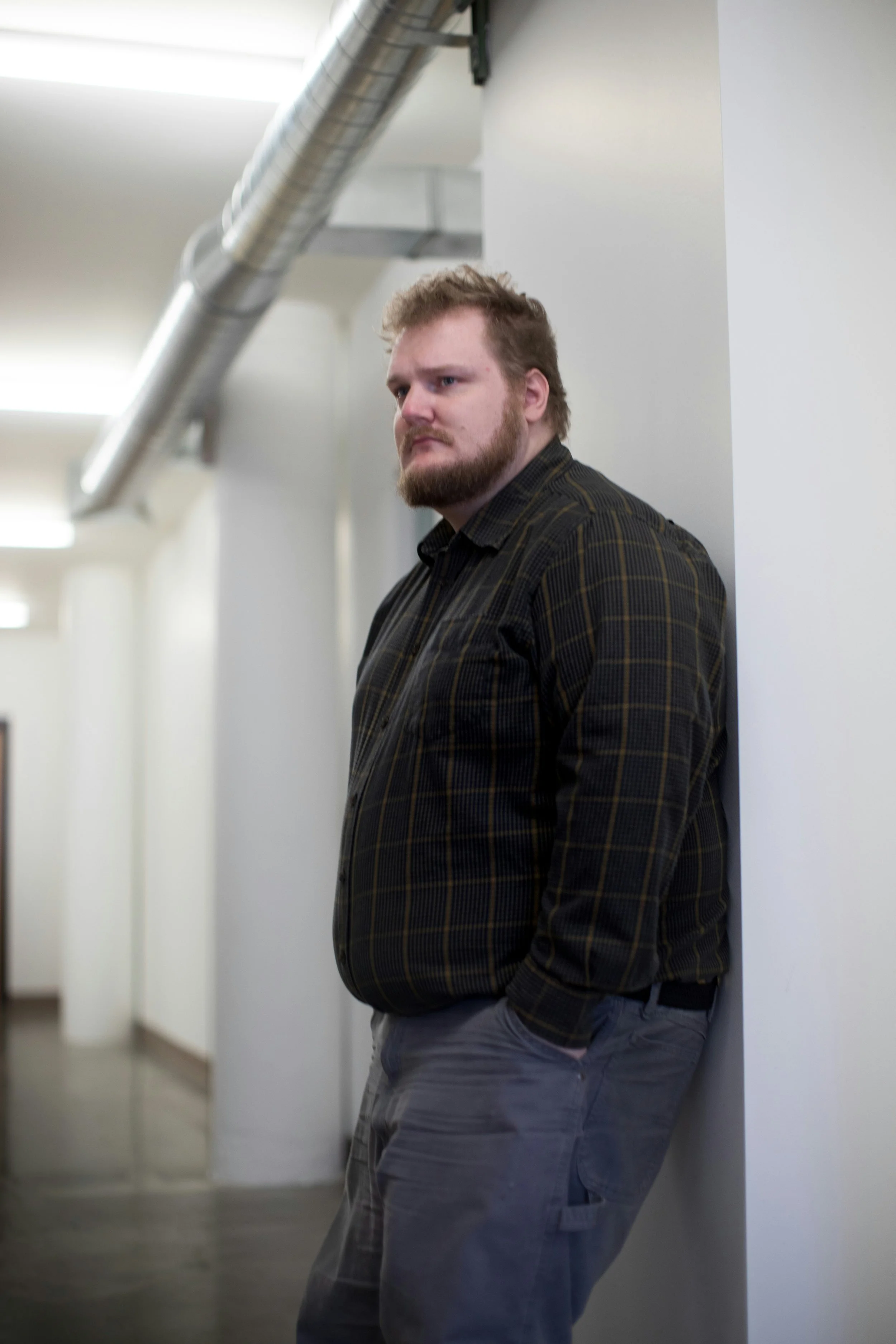 A man with a beard and casual clothing leaning against a white wall in an interior space with white walls and ceiling fixtures.