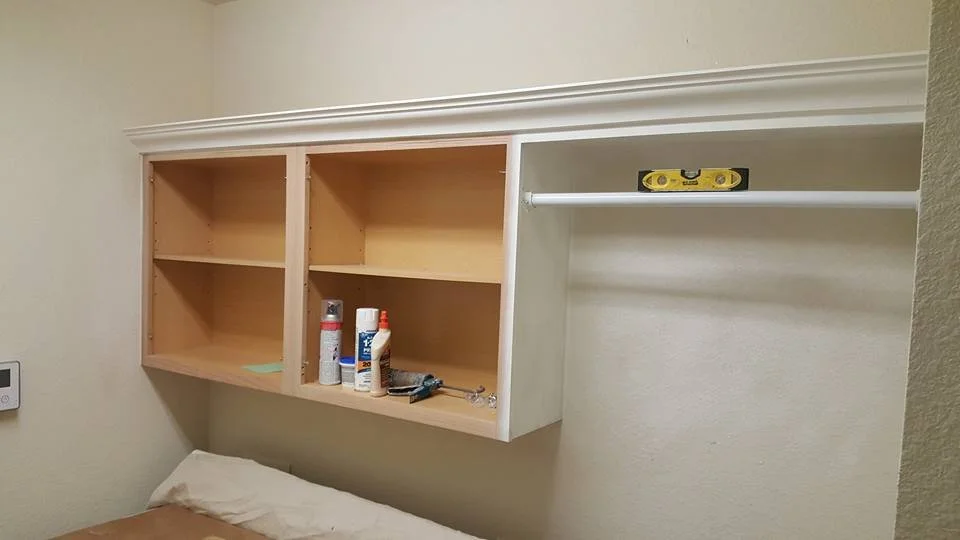 Wooden wall cabinet with open shelves mounted above a bed, containing bottles of cleaning supplies and small tools, in a bedroom.