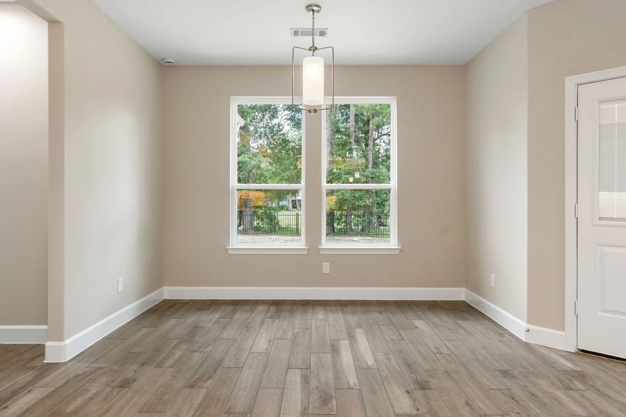 Empty dining room with two large windows showing trees outside, a hanging light fixture, beige walls, white baseboards, and wood-look tile flooring.