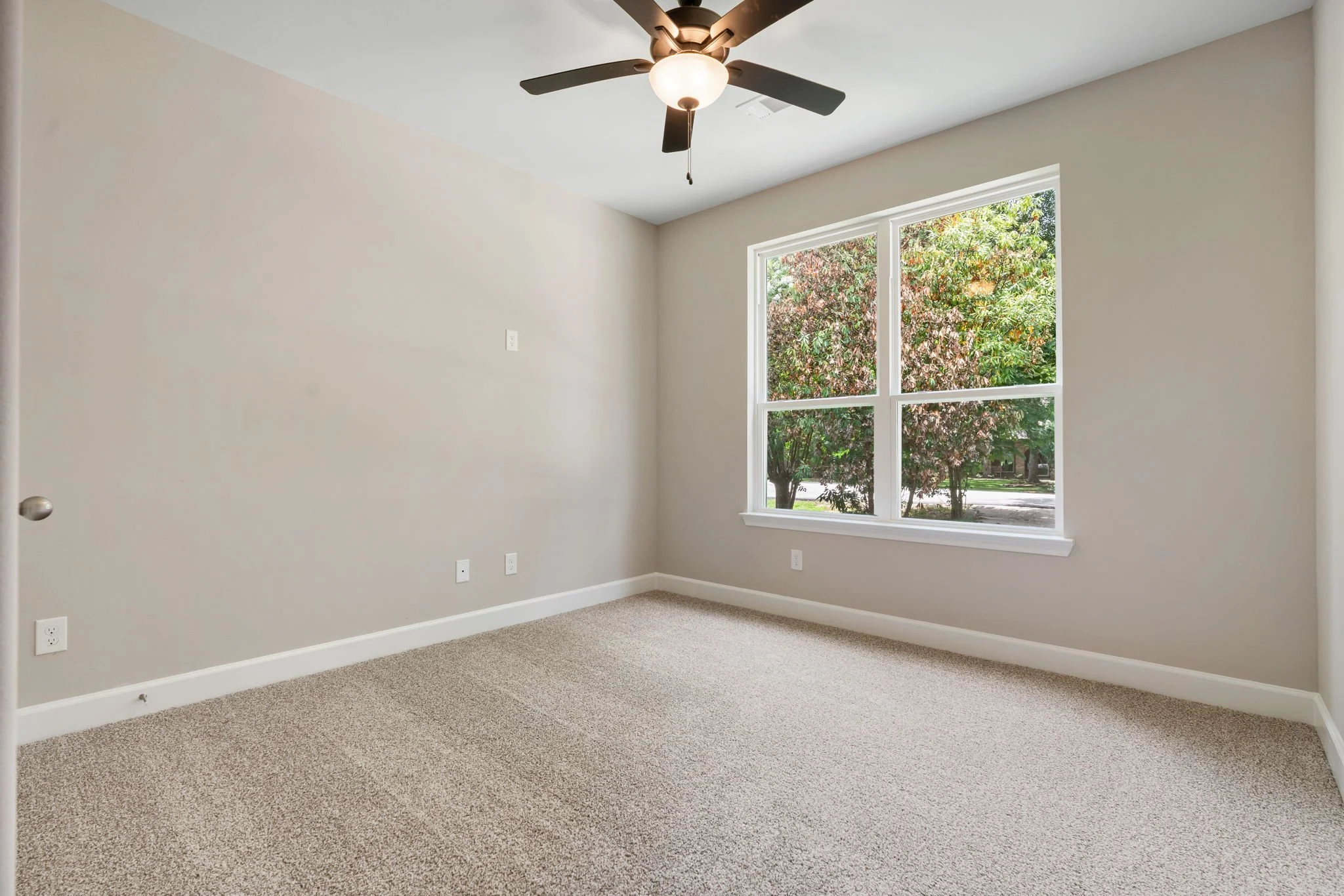 Empty room with beige carpet, cream walls, large window showing trees outside, ceiling fan with light.