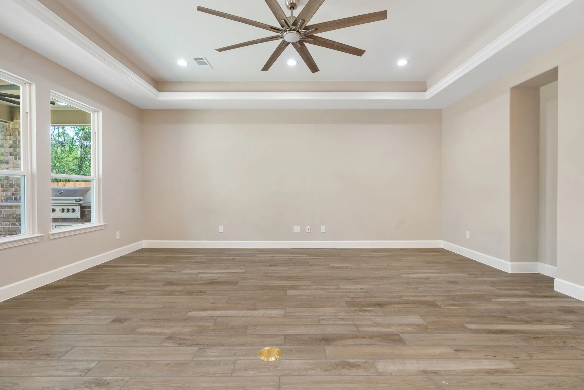 Empty living room with light beige walls, hardwood floors, white baseboards, and ceiling with crown molding. Features three windows and ceiling fan with wooden blades.