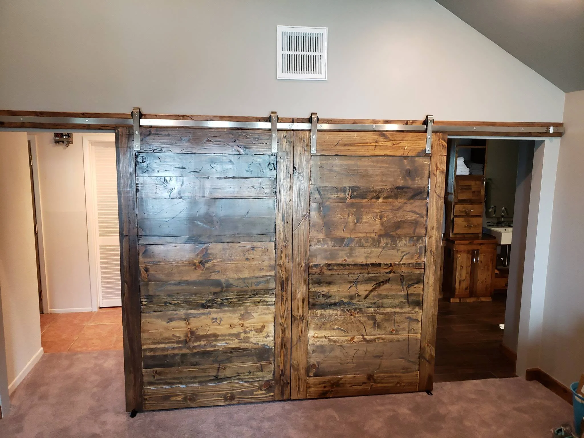 A wooden sliding barn door partially open revealing a bathroom with a small sink and wooden cabinetry in a room with beige walls and carpeted flooring.