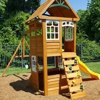 Wooden playground playset with a slide and climbing ladder on a sandy surface, surrounded by greenery.