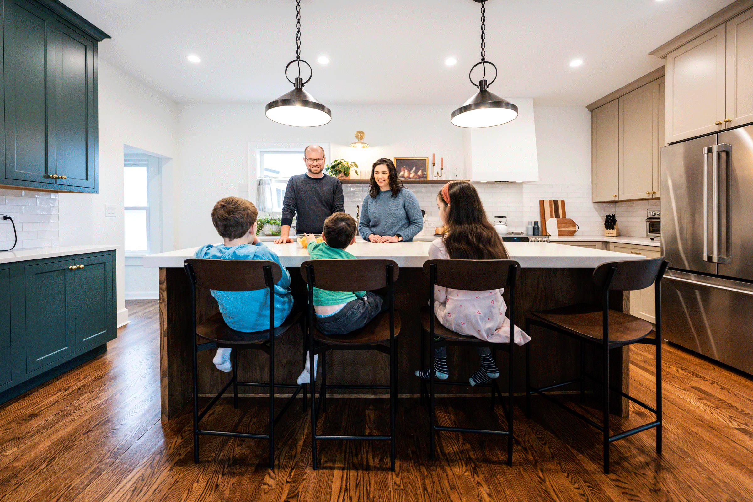 A family gathering around a kitchen island with parents and three children, engaged in conversation in a modern kitchen with white cabinets, wooden floors, and minimal decor.