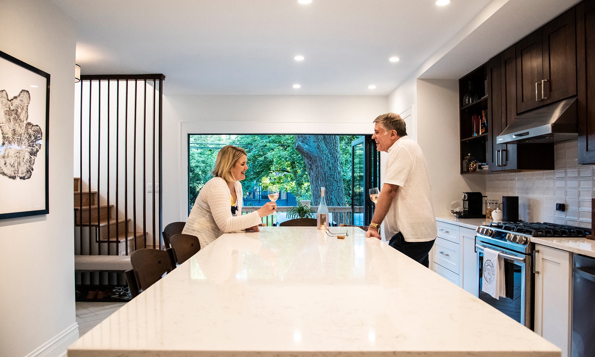 A man and woman having drinks and talking in a kitchen with white cabinets, a large island, and a window overlooking a green backyard with a large tree.