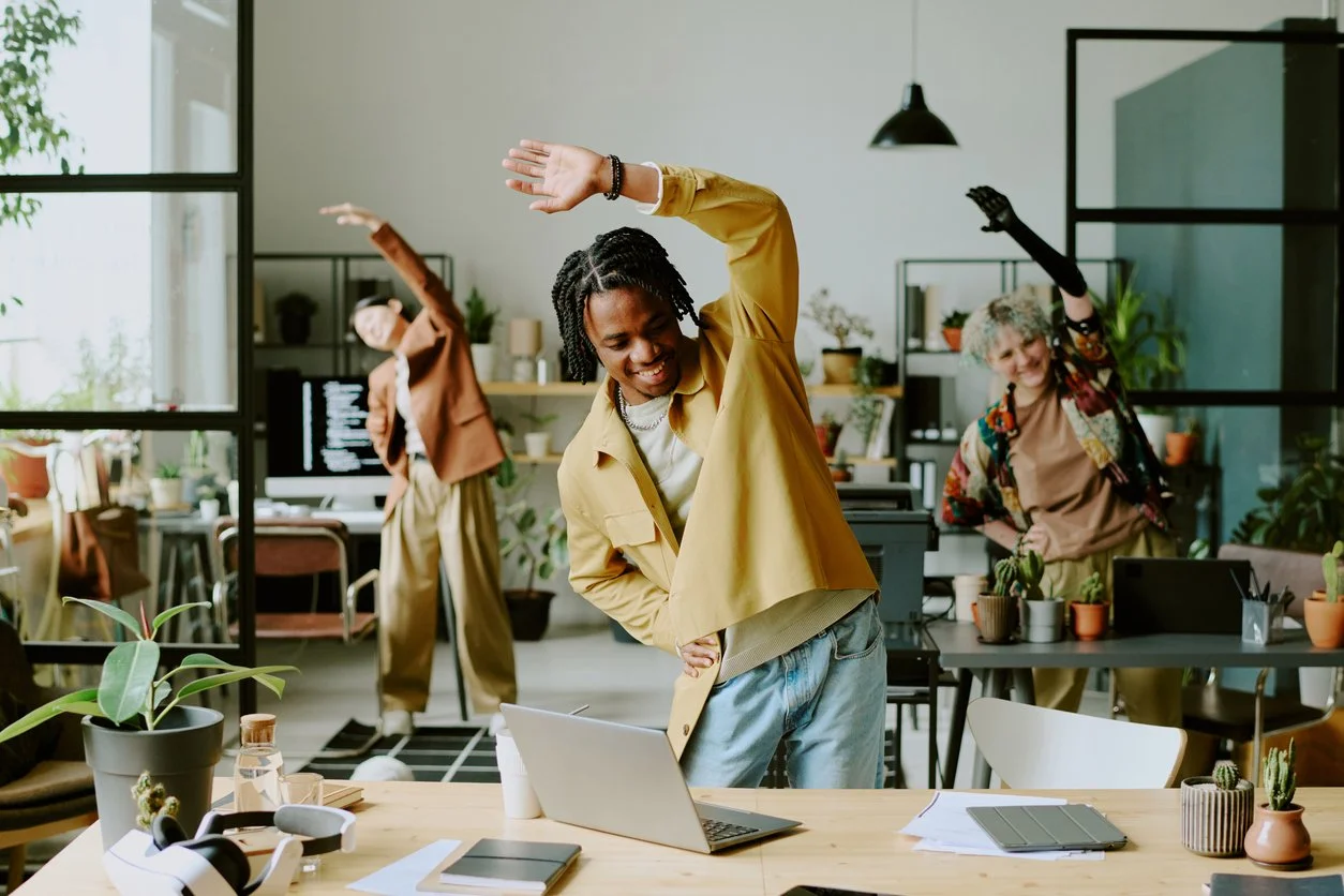 A woman in a mustard-colored top and brown pants practicing Pilates in her living room, in front of a laptop on the floor.