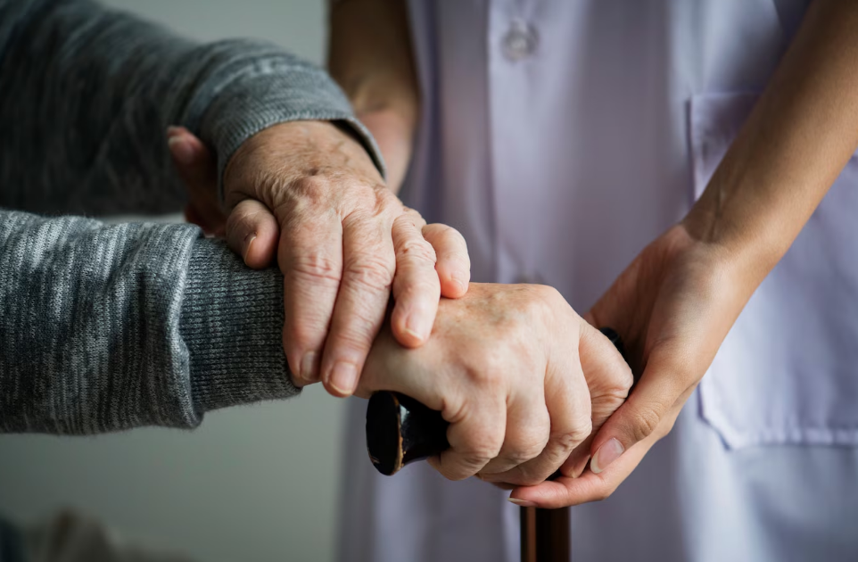 Close-up of an elderly person's hand holding a cane, while a caregiver's hand gently supports it, symbolizing assistance and care.