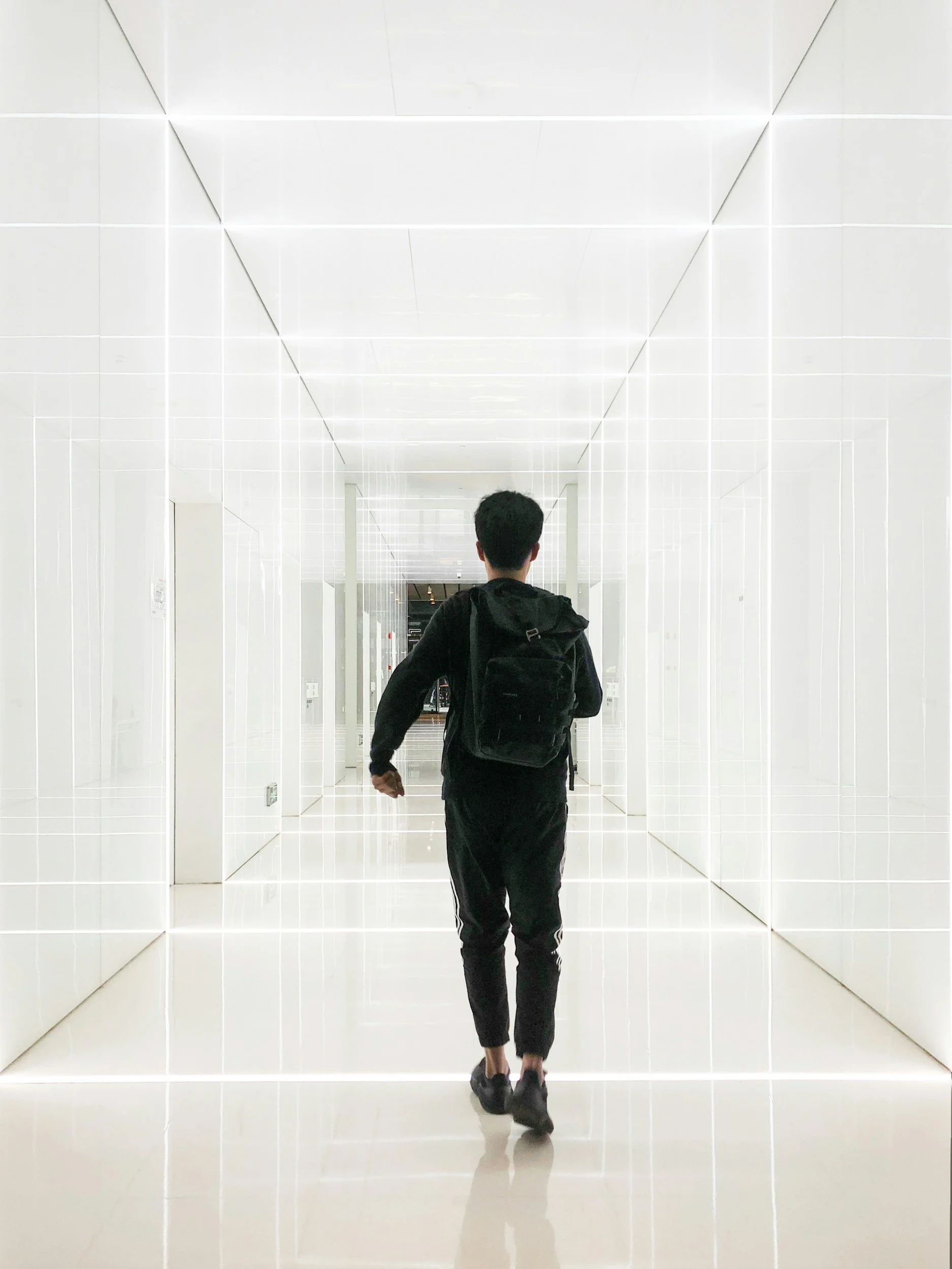 A person walking through a sleek, brightly lit, white corridor with geometric wall and ceiling lines, carrying a black backpack, viewed from behind.