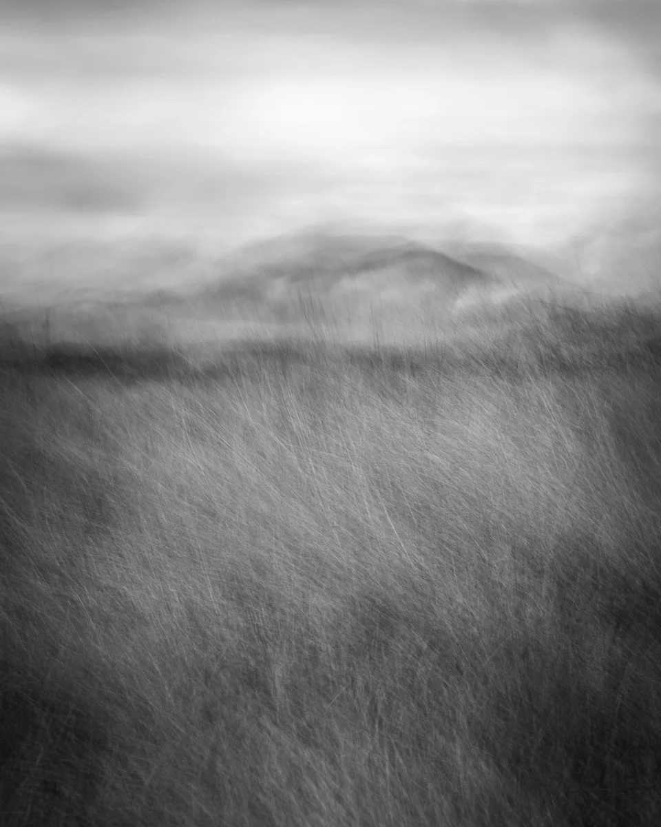 An ICM impressionist black and white landscape with grassy terrain and a distant hill under a cloudy sky.
