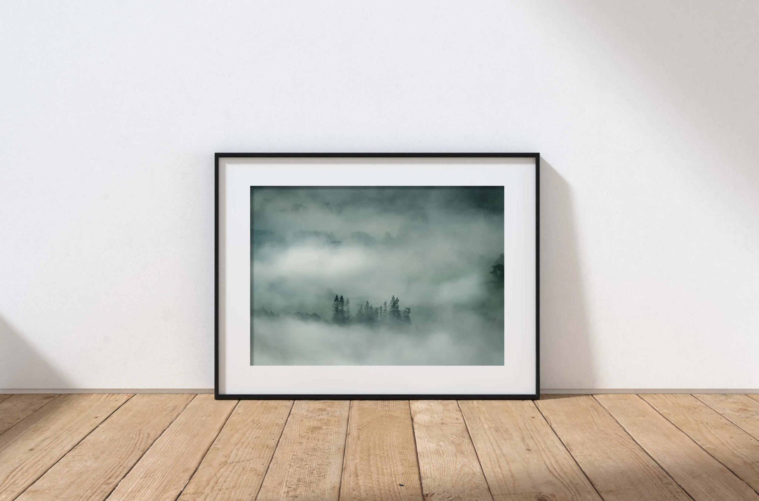 Framed landscape photograph of misty mountains and pine trees, leaning against a white wall on a wooden floor.